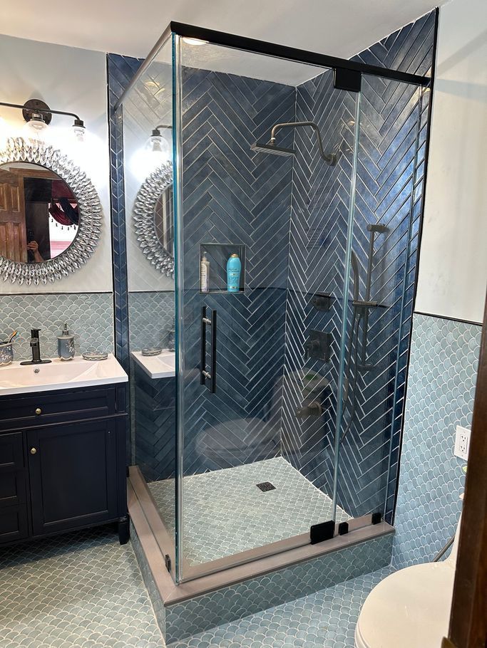 Modern bathroom with a glass-enclosed shower, blue herringbone tile, and dark vanity with round mirrors