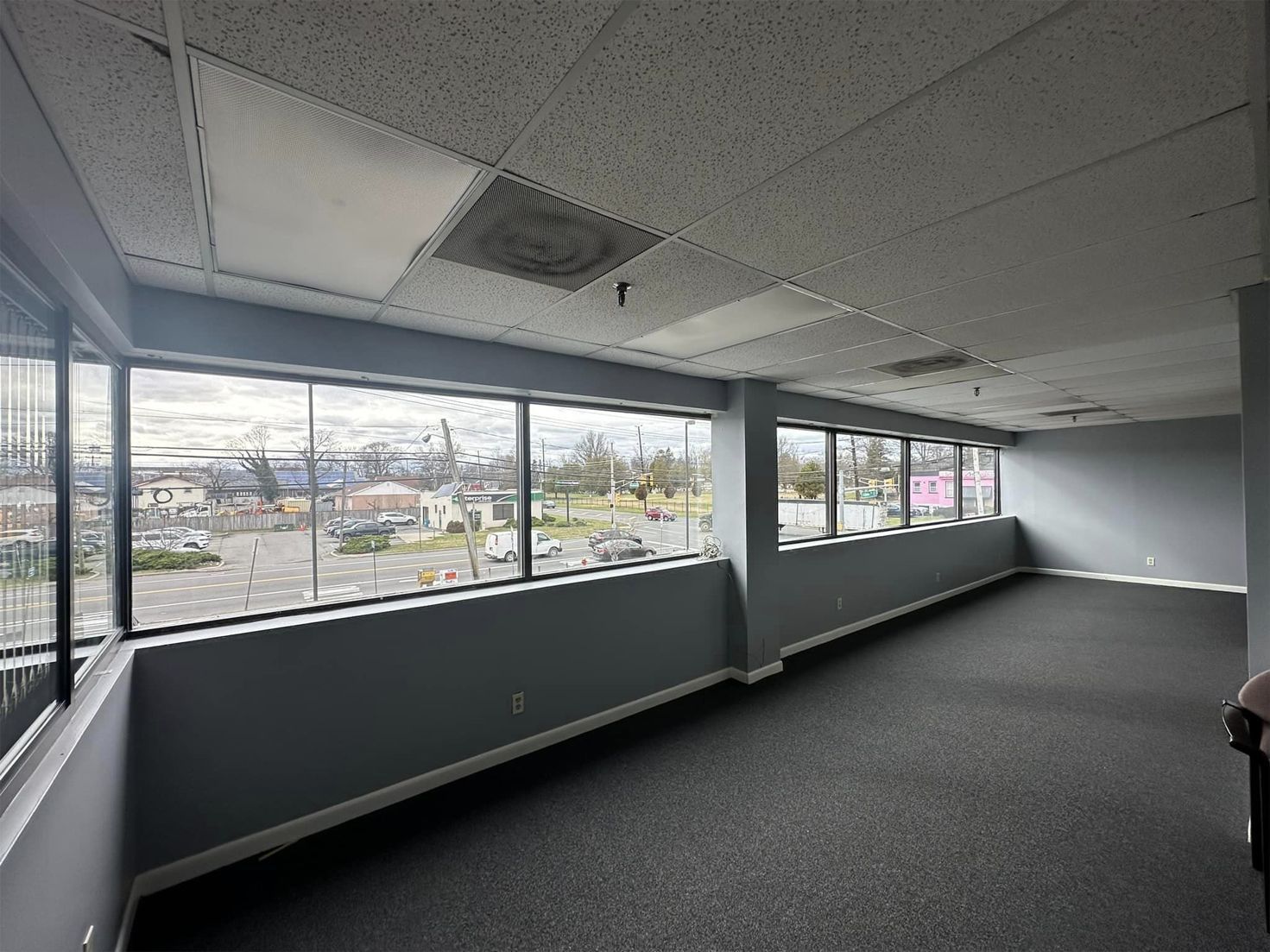 Empty office room with large windows, gray carpet, and drop ceiling tiles overlooking a city view