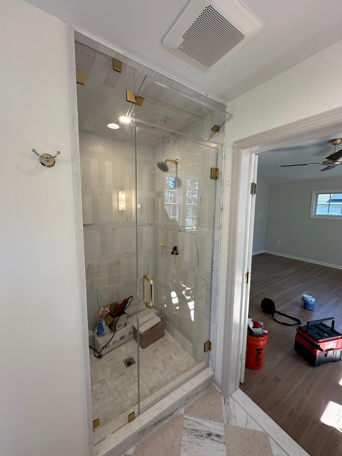Bathroom under renovation with glass shower enclosure beside an open doorway to a room with wood flooring.