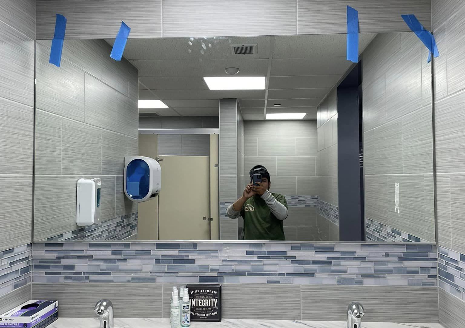 Mirror selfie in a tiled restroom with blue-gray accents and sinks visible below.