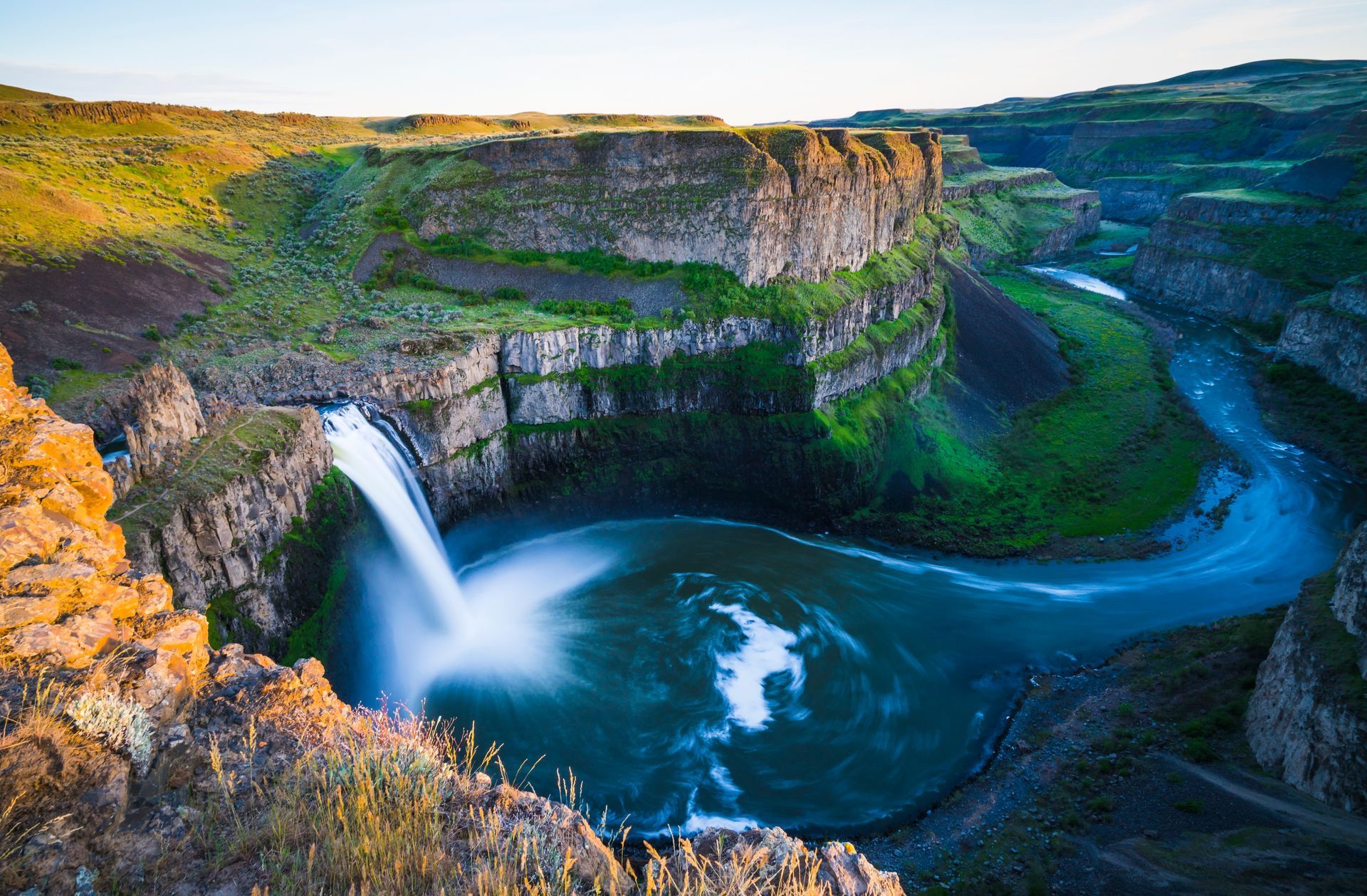 Waterfall plunging into a turquoise canyon river at sunset, with green cliffs and rocky gorge.