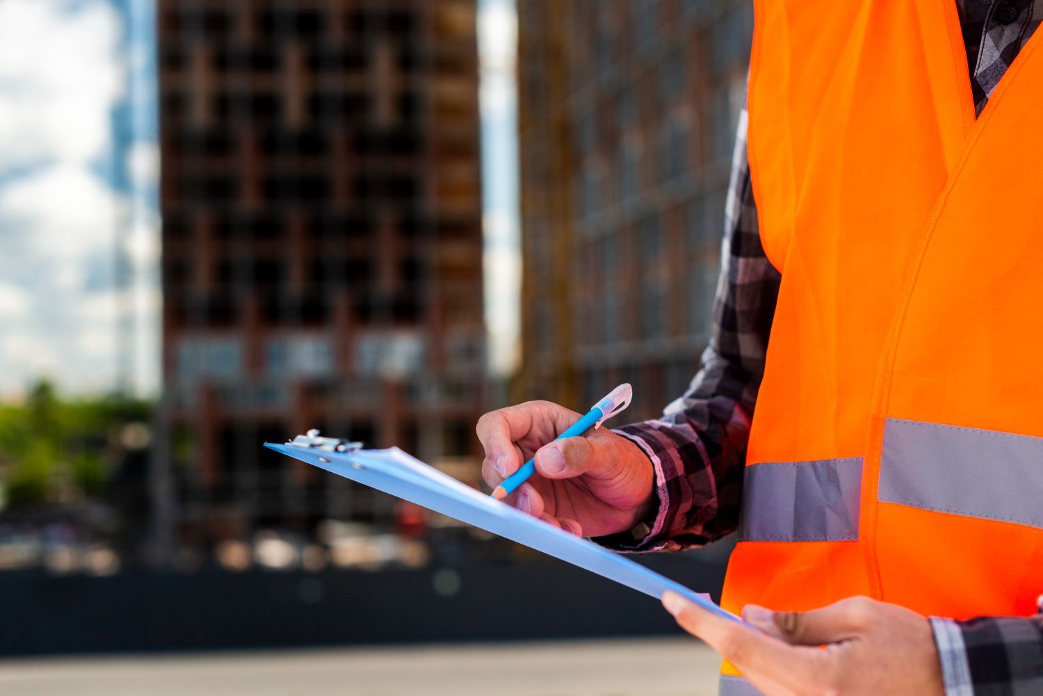 Construction worker in orange vest, holding clipboard and pen, building in background.