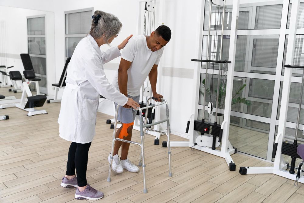 Therapist assisting a patient using a walker in a physical therapy gym. Patient wears a knee brace.