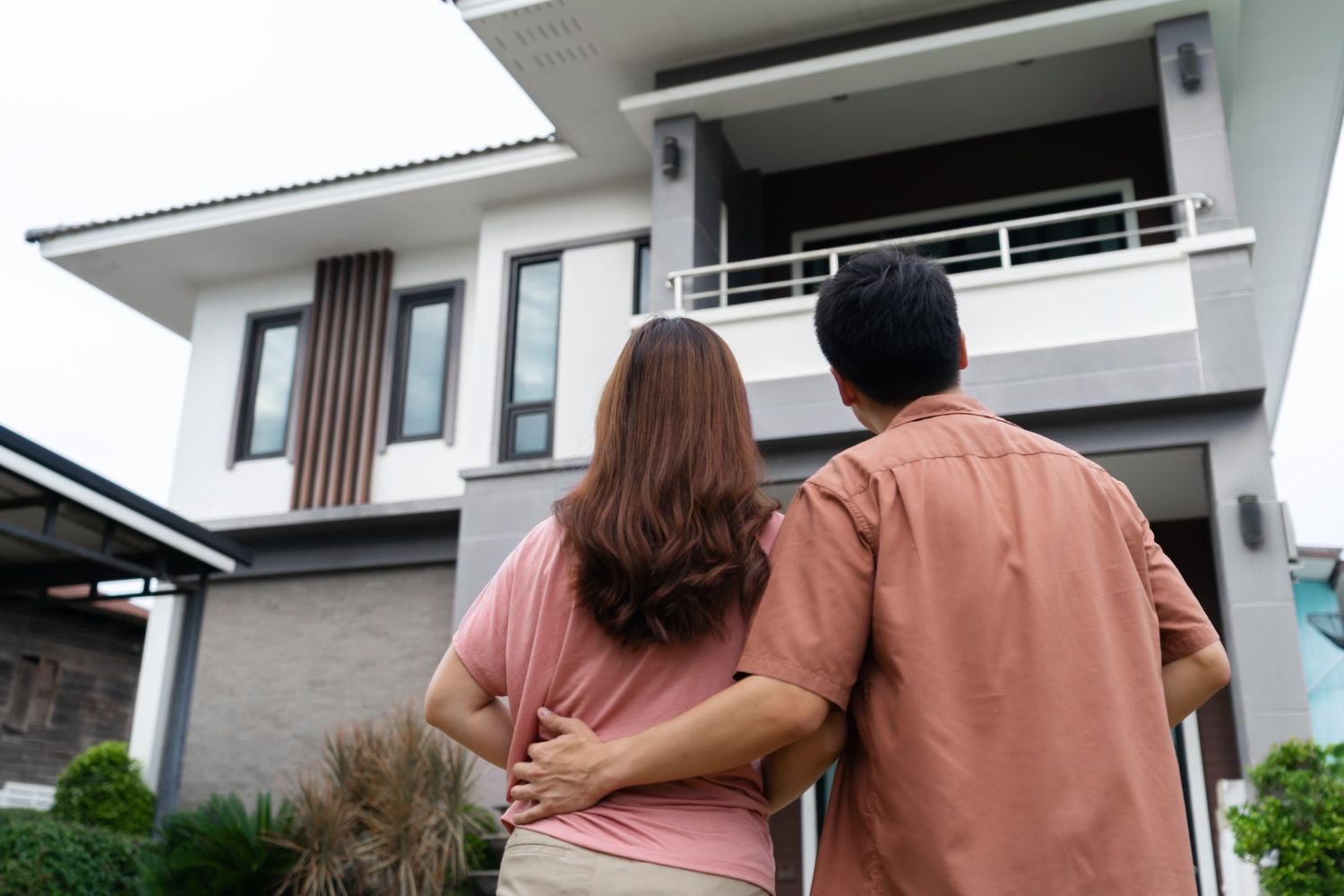 Couple gazing at a modern two-story house, arm in arm. Exterior shot, cloudy sky, neutral colors.