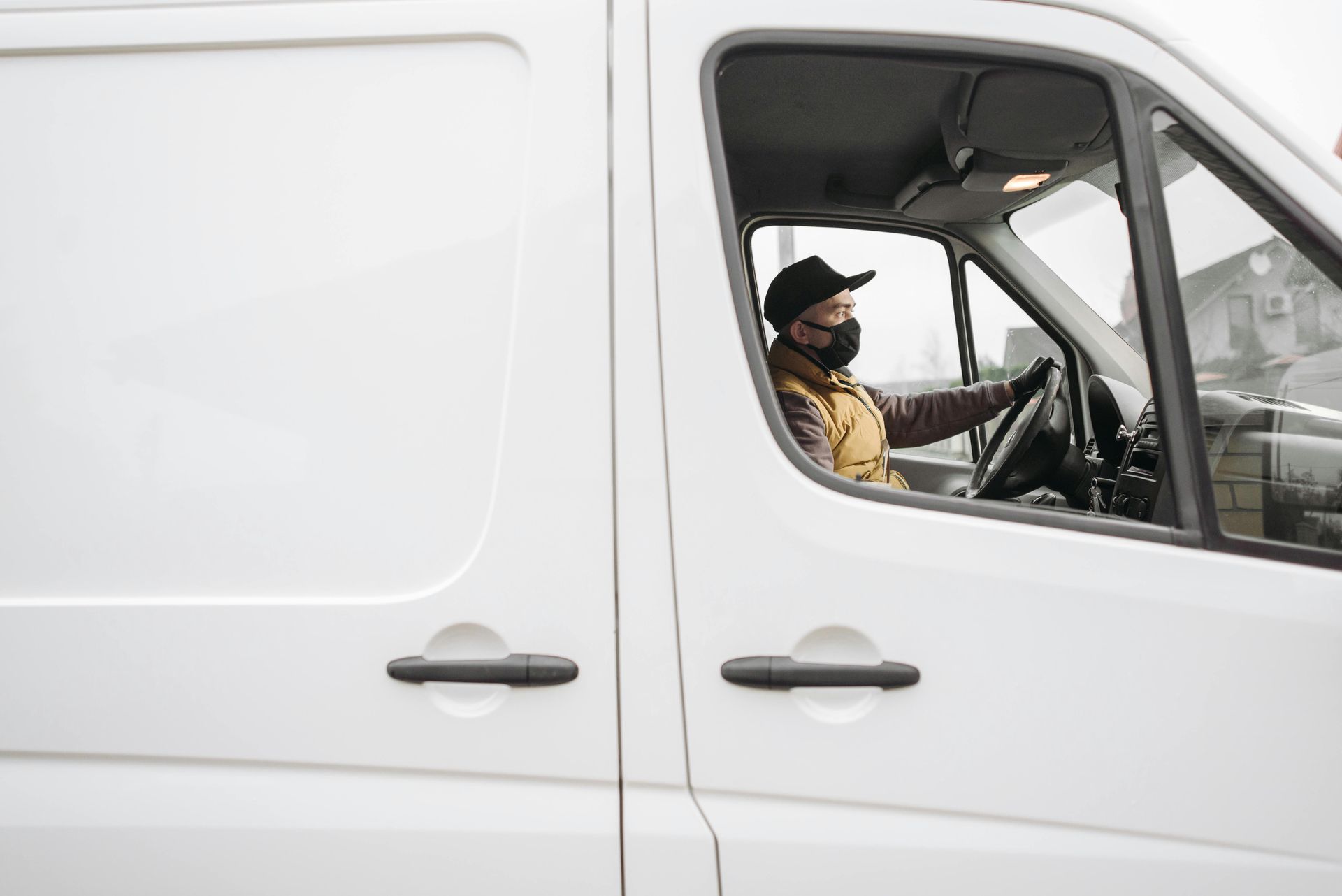 A person wearing a face mask and cap drives a white van.
