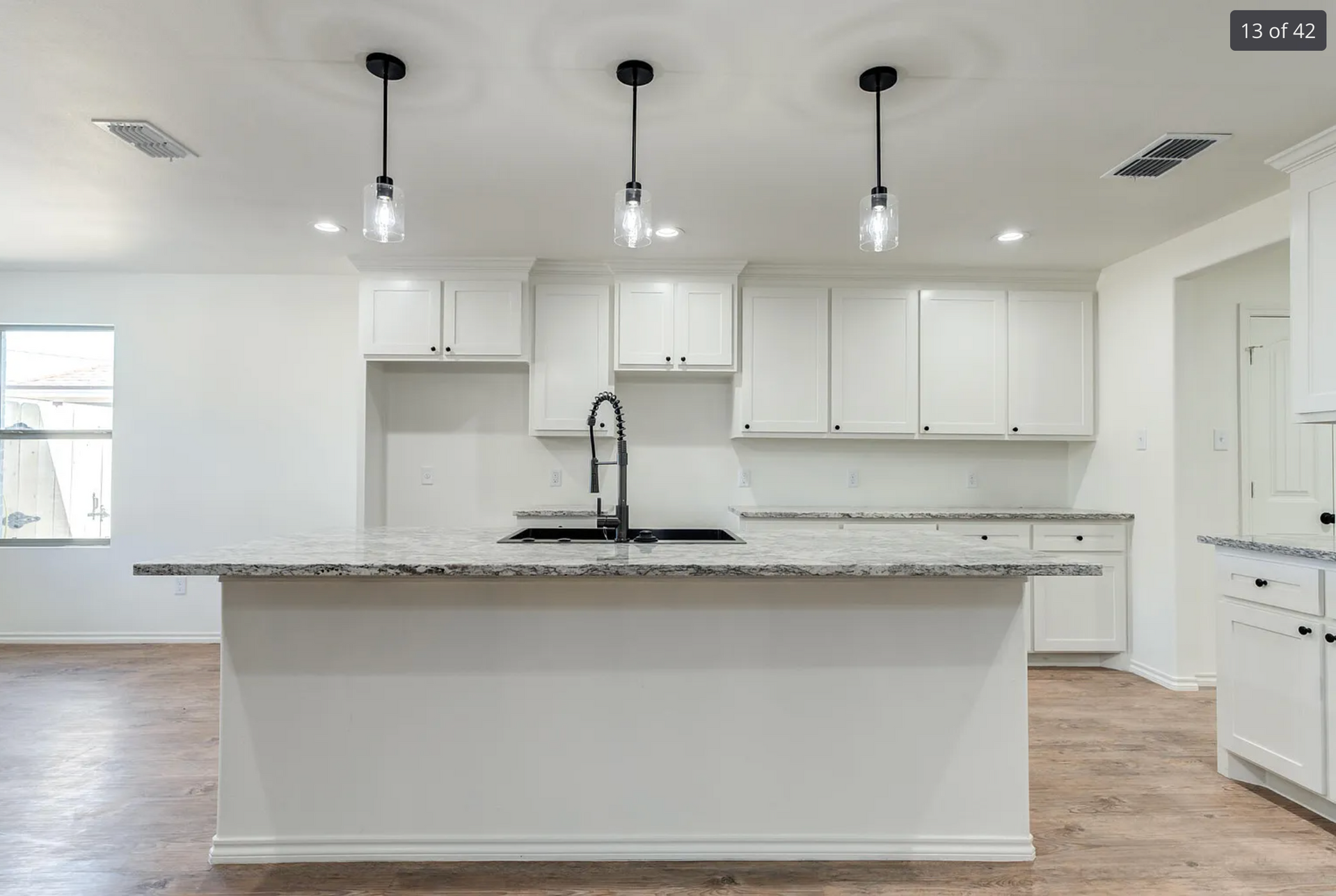 Bright kitchen with white cabinets, granite countertops, and a large island. Three pendant lights hang above.