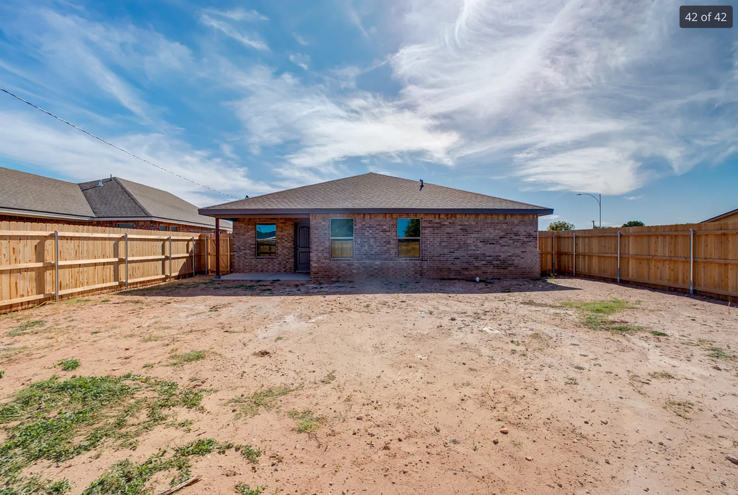 Backyard of a brick house with wooden fence, blue sky, and barren ground.