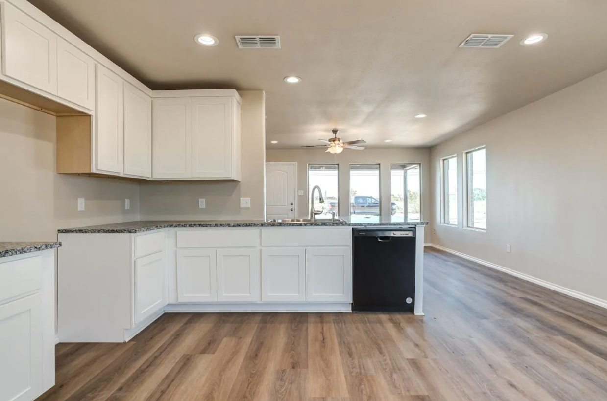 Bright empty kitchen and living area with white cabinets, wood floors, and large windows.