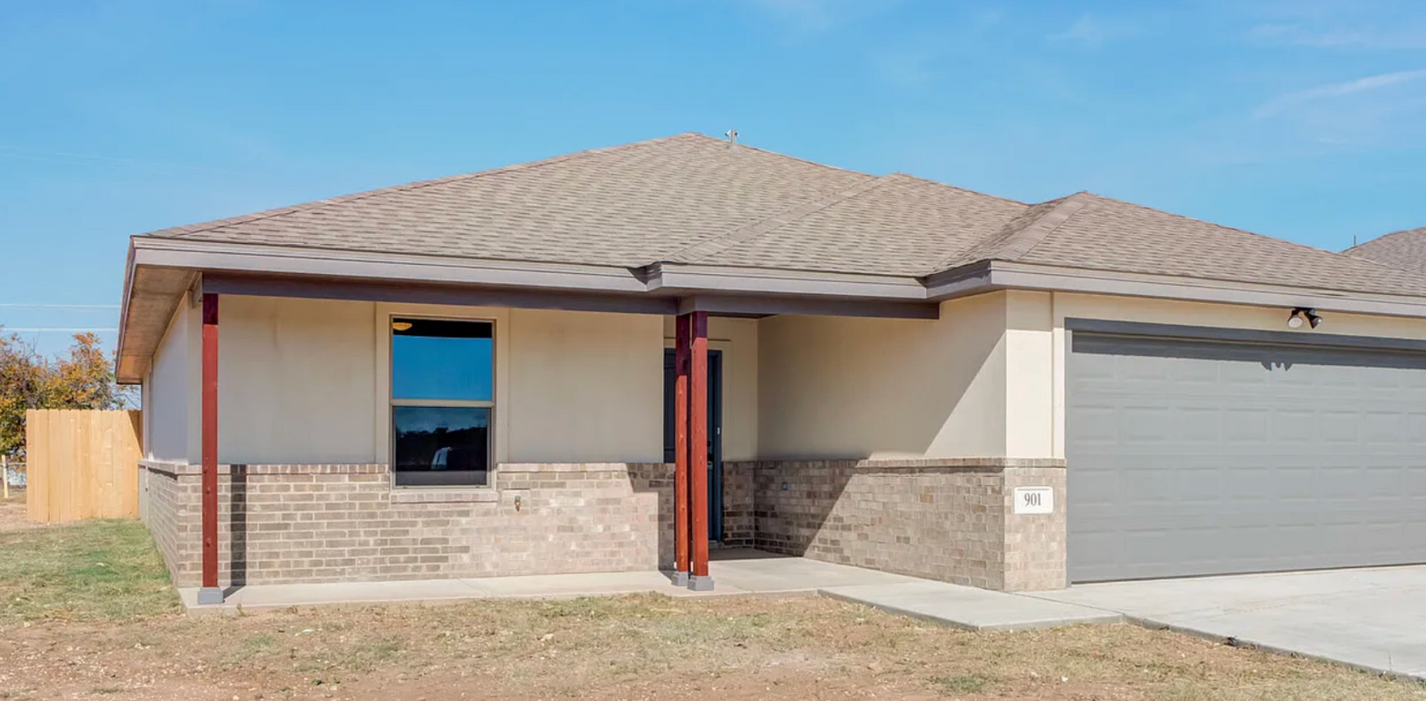 Tan house with gray garage door, brown roof, and brick facade on a sunny day.