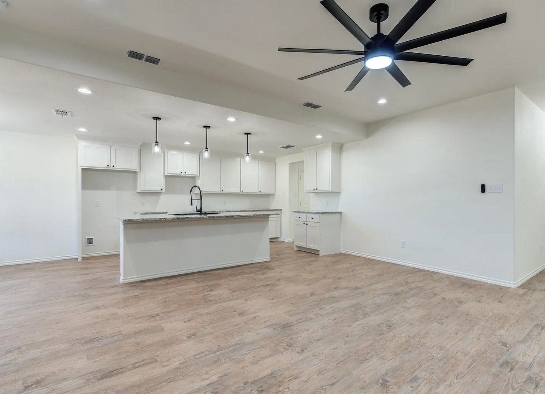 Empty modern kitchen and living area with white cabinets, island, wood floors, and black ceiling fan.