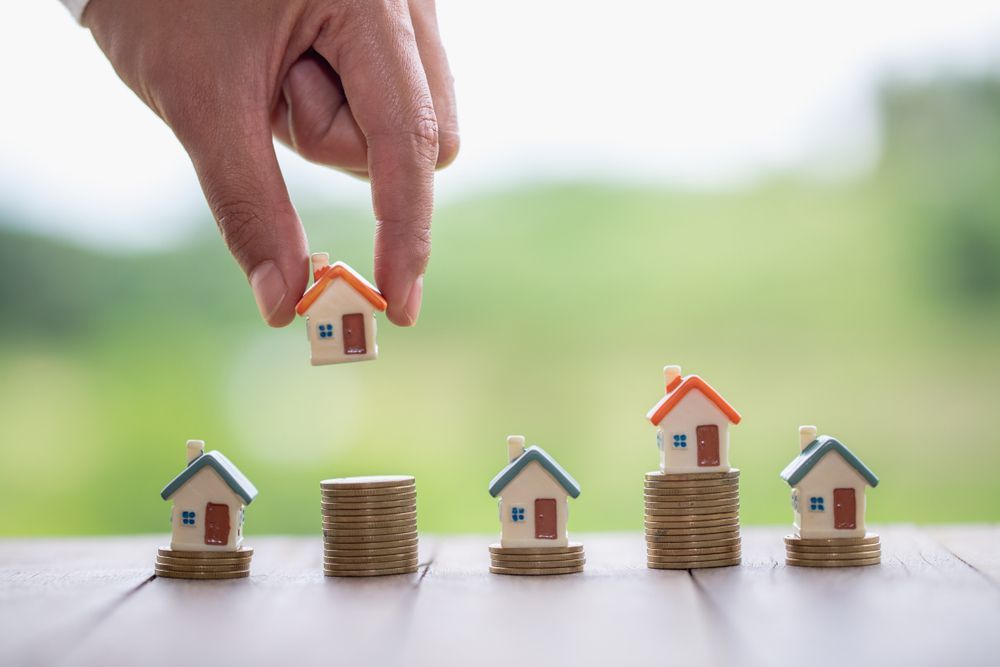 A person is putting a small house on top of a pile of coins.