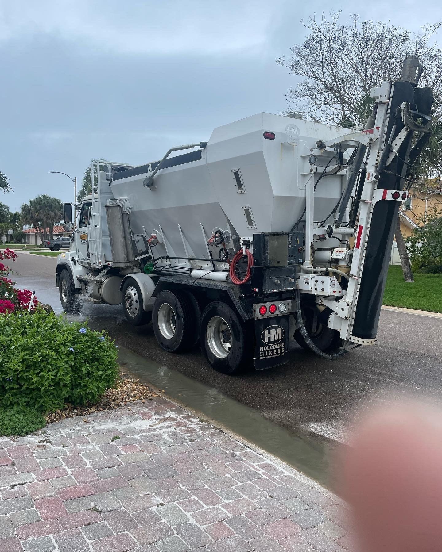 White dump truck driving on a wet road beside a sidewalk and bushes.