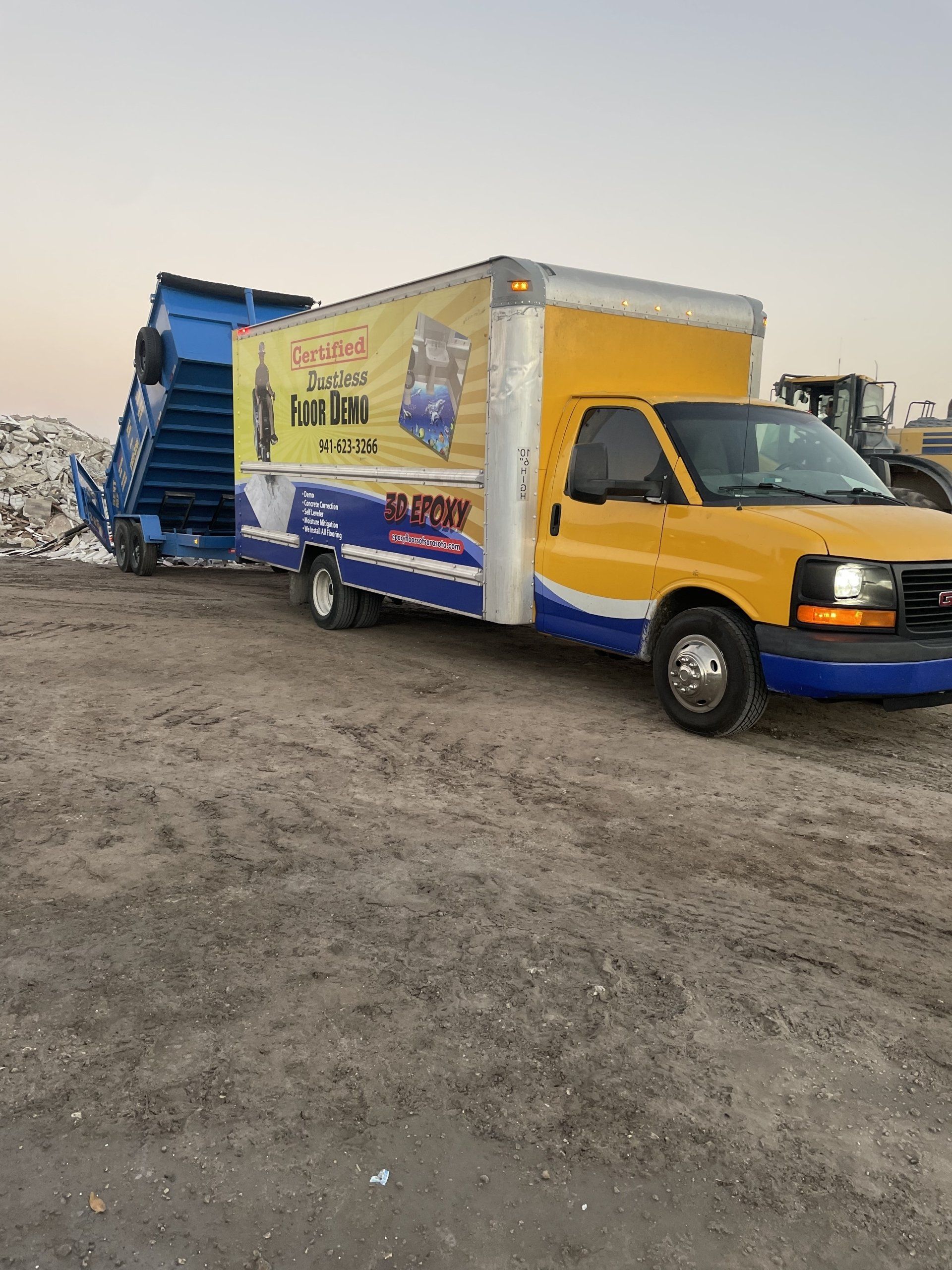 Yellow box truck with a blue cargo ramp on a dirt lot at sunset