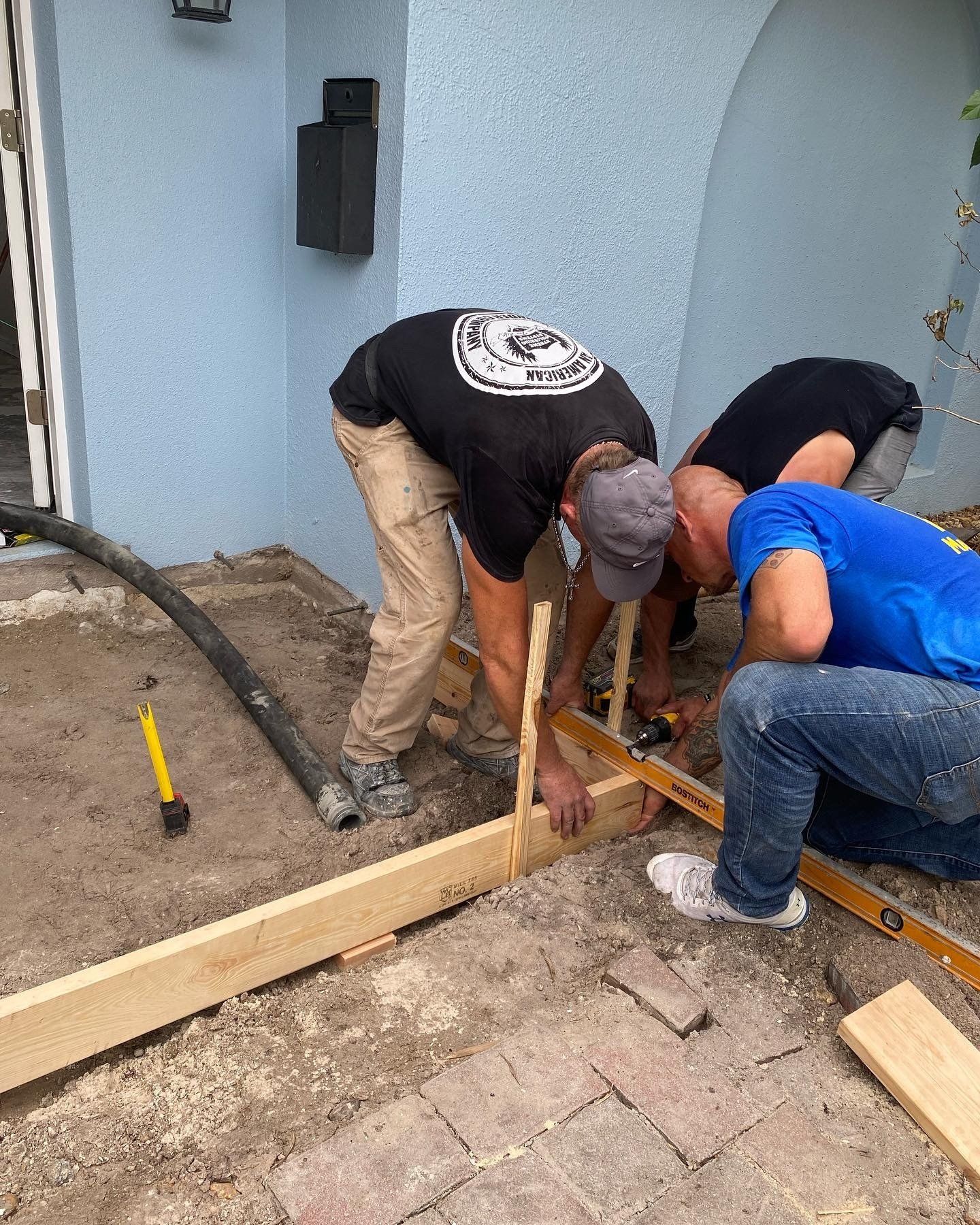 Three workers installing wooden framing beside a house, crouched over a sidewalk area.