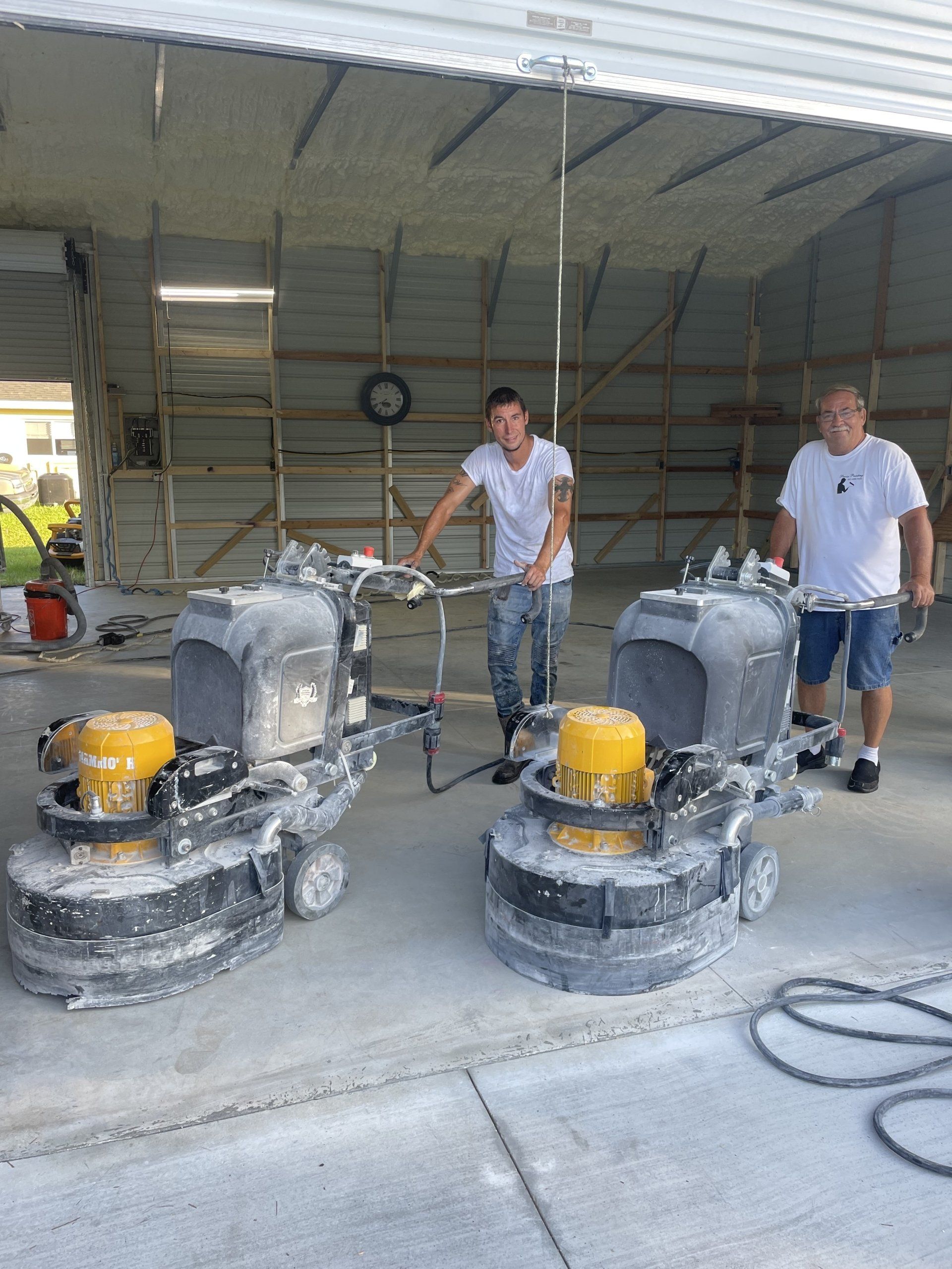 Two workers operate floor grinders in a large workshop, with dust-covered concrete and equipment around them.