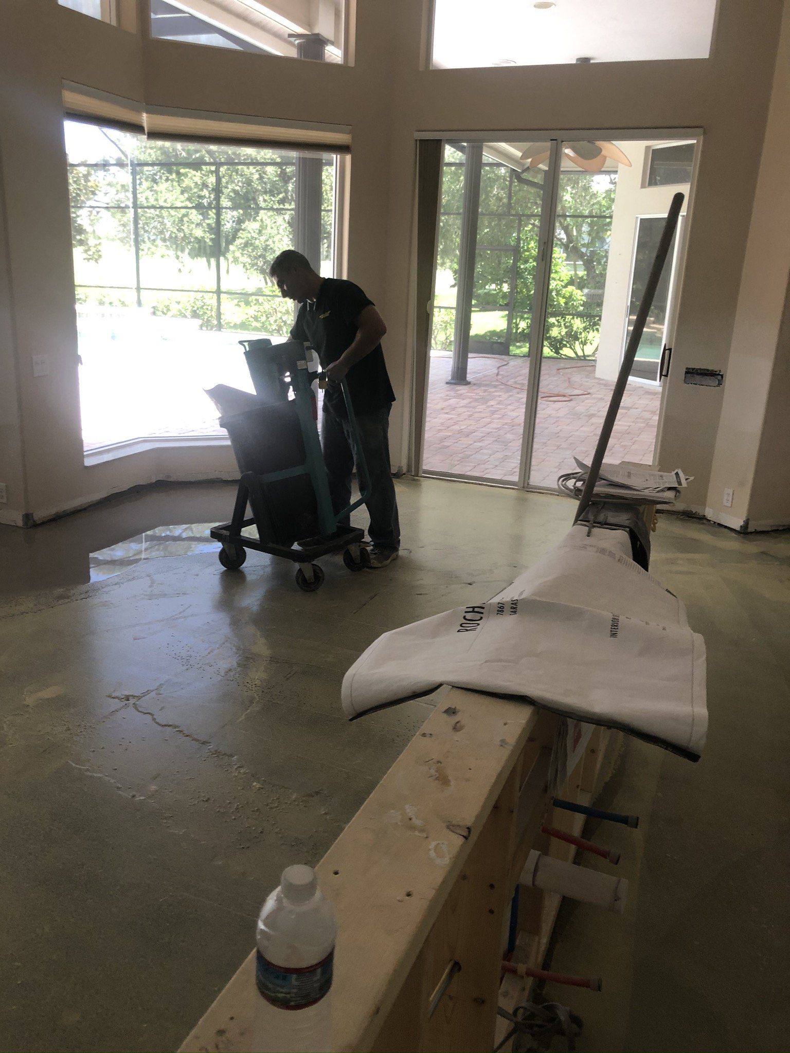 Worker using a floor machine in a bright, unfinished room with large windows and scattered construction materials
