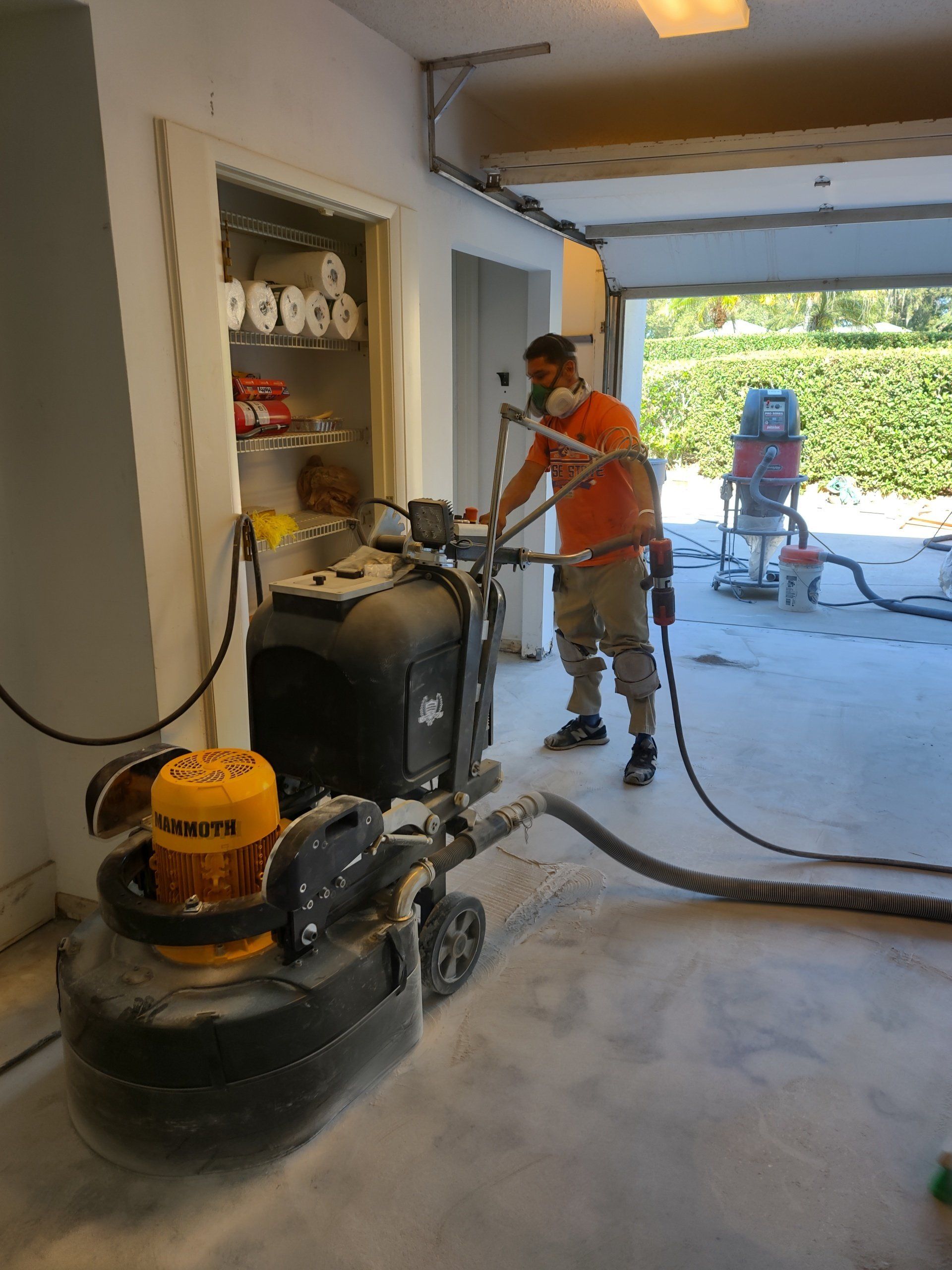 Worker using a floor grinder in a garage during concrete resurfacing, with hoses and tools nearby