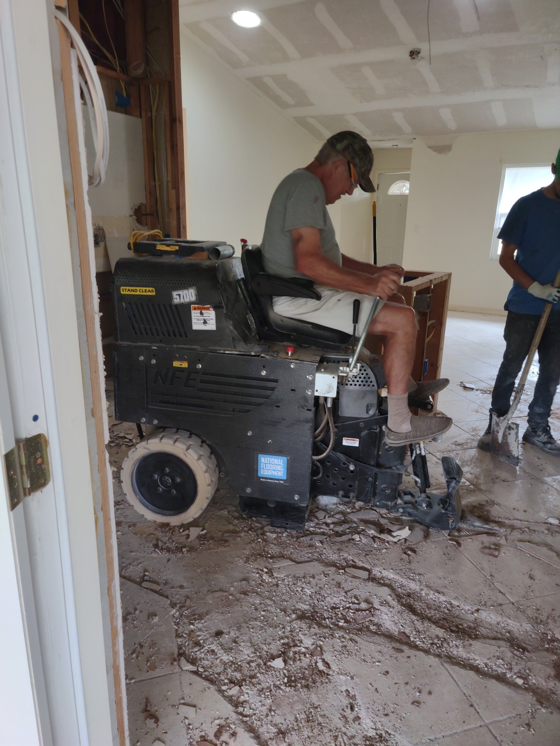 Man on a black skid steer inside a room, with mud scattered across the floor; another person stands nearby.