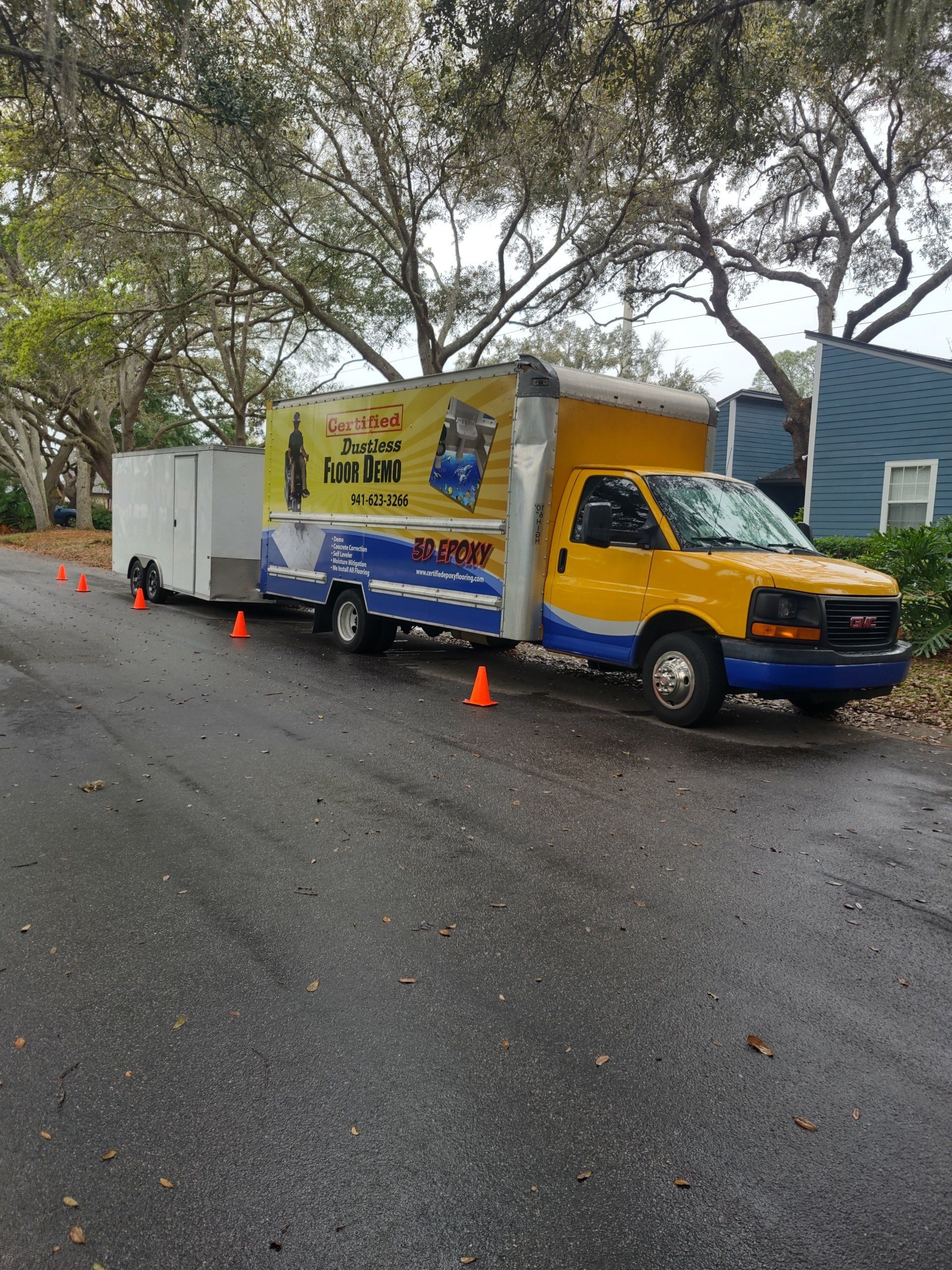 Yellow box truck and white trailer parked on a wet roadside with orange cones under trees