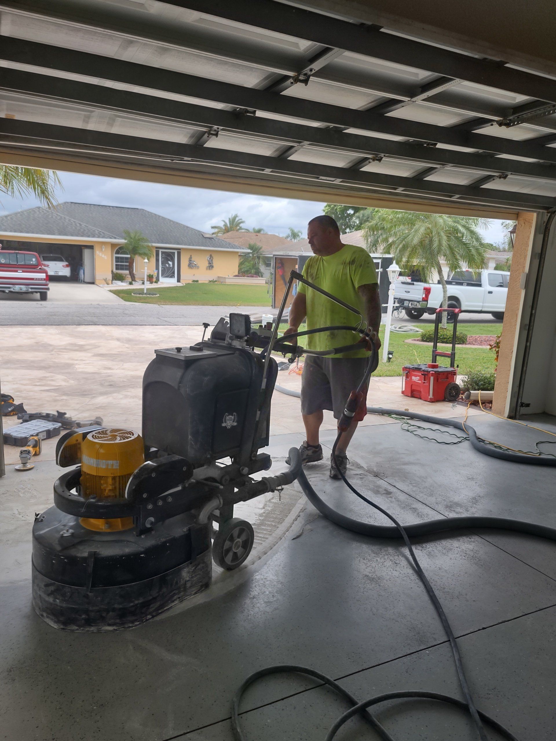 Worker pressure washes a garage floor with a floor buffer, hose trailing across the concrete.