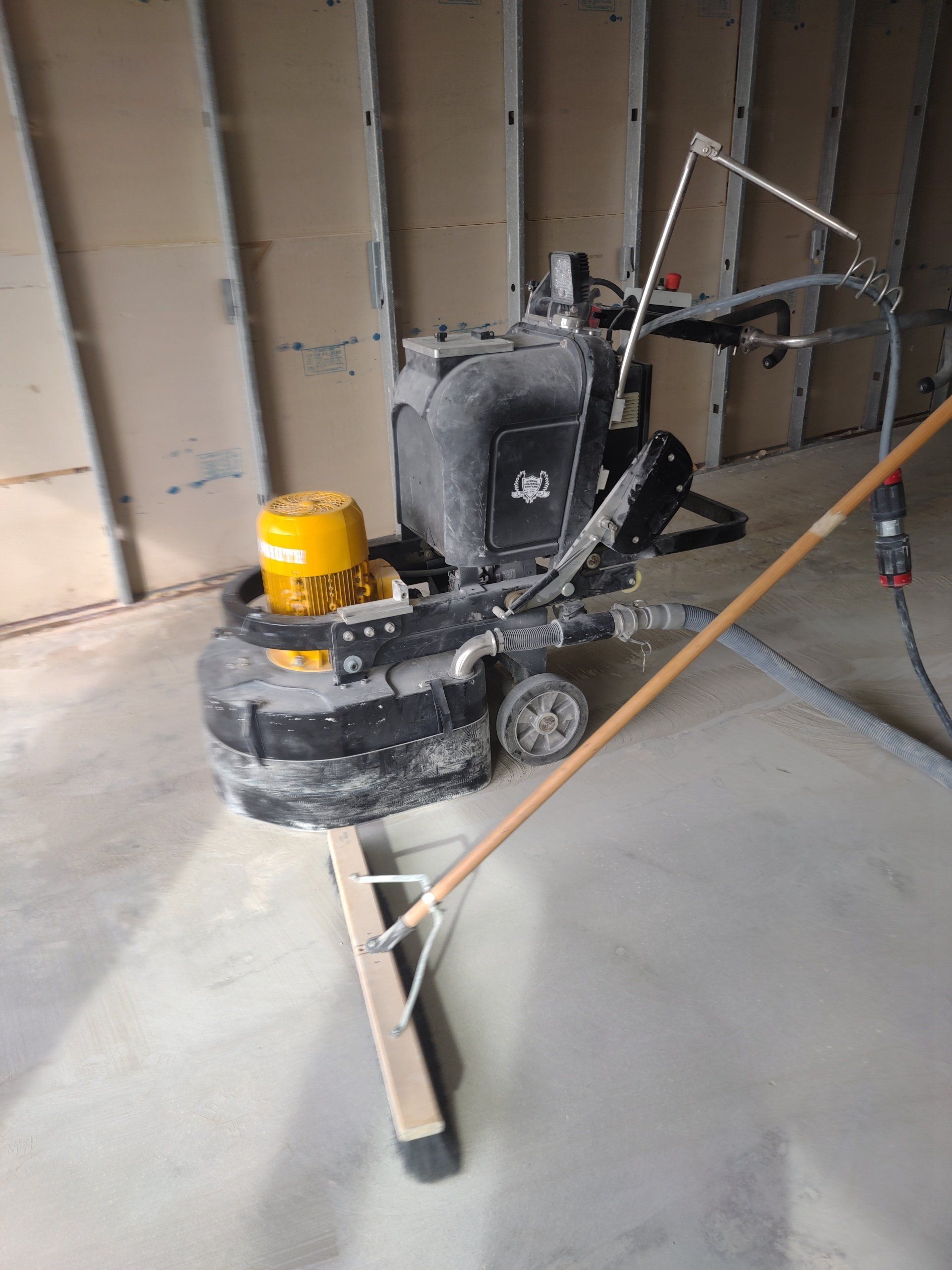 Black and yellow floor grinder on concrete inside an unfinished room with exposed wall studs