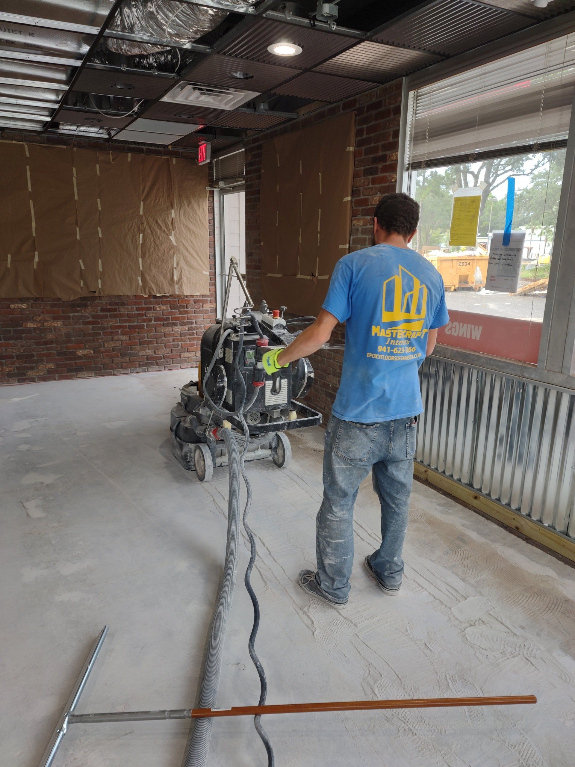 Worker operating a floor grinder in an unfinished garage with exposed brick and open doorway.