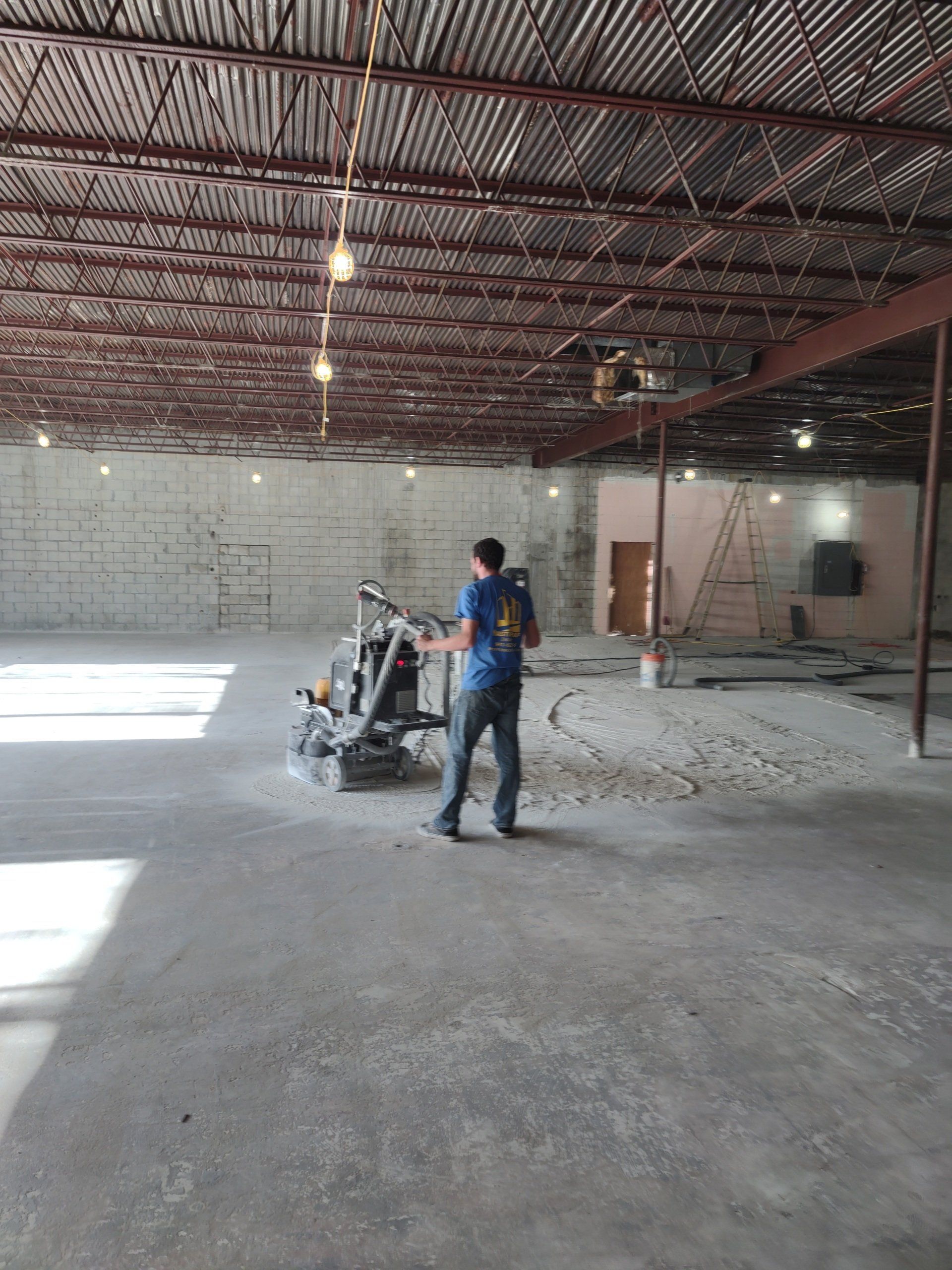 Worker operating a concrete grinder inside a large unfinished industrial building with exposed ceiling beams.