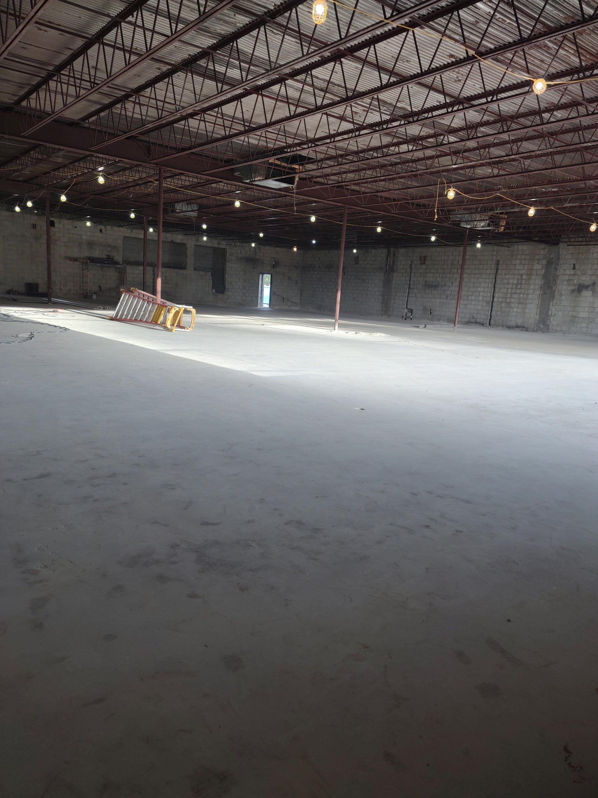 Empty indoor warehouse with a metal roof and concrete floor, lit by a few hanging lights