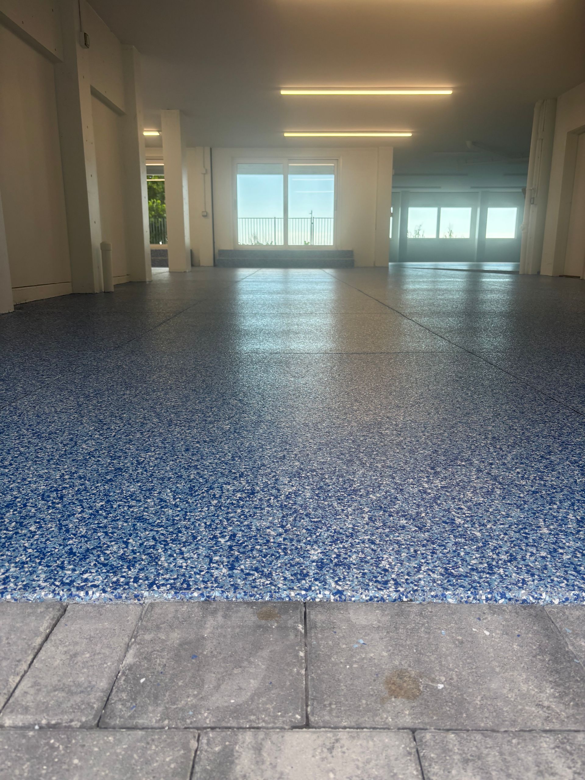 Empty indoor pool area with blue-speckled floor, tiled edge, and large windows at the far end
