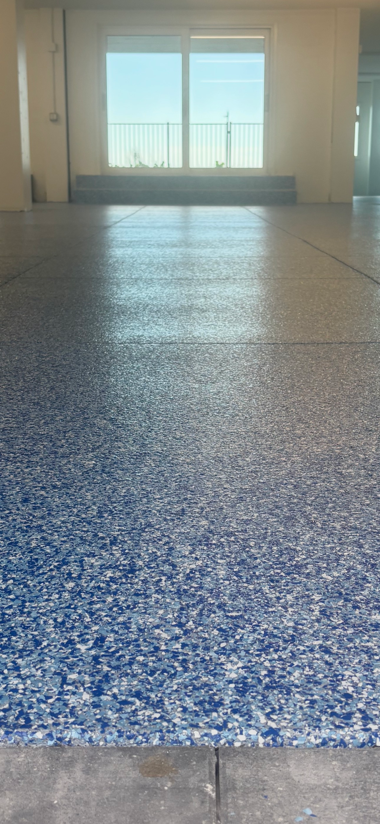 Blue-speckled floor in a bright empty room with large windows and sunlight reflecting off the surface