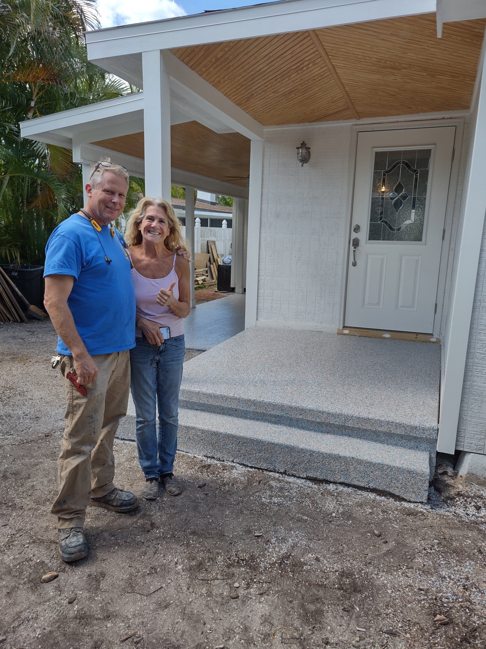Two people standing on a front porch of a small white house, smiling at the camera