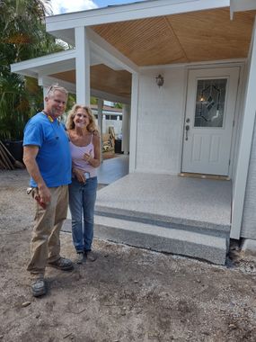 Two people standing on a front porch of a small white house, smiling at the camera