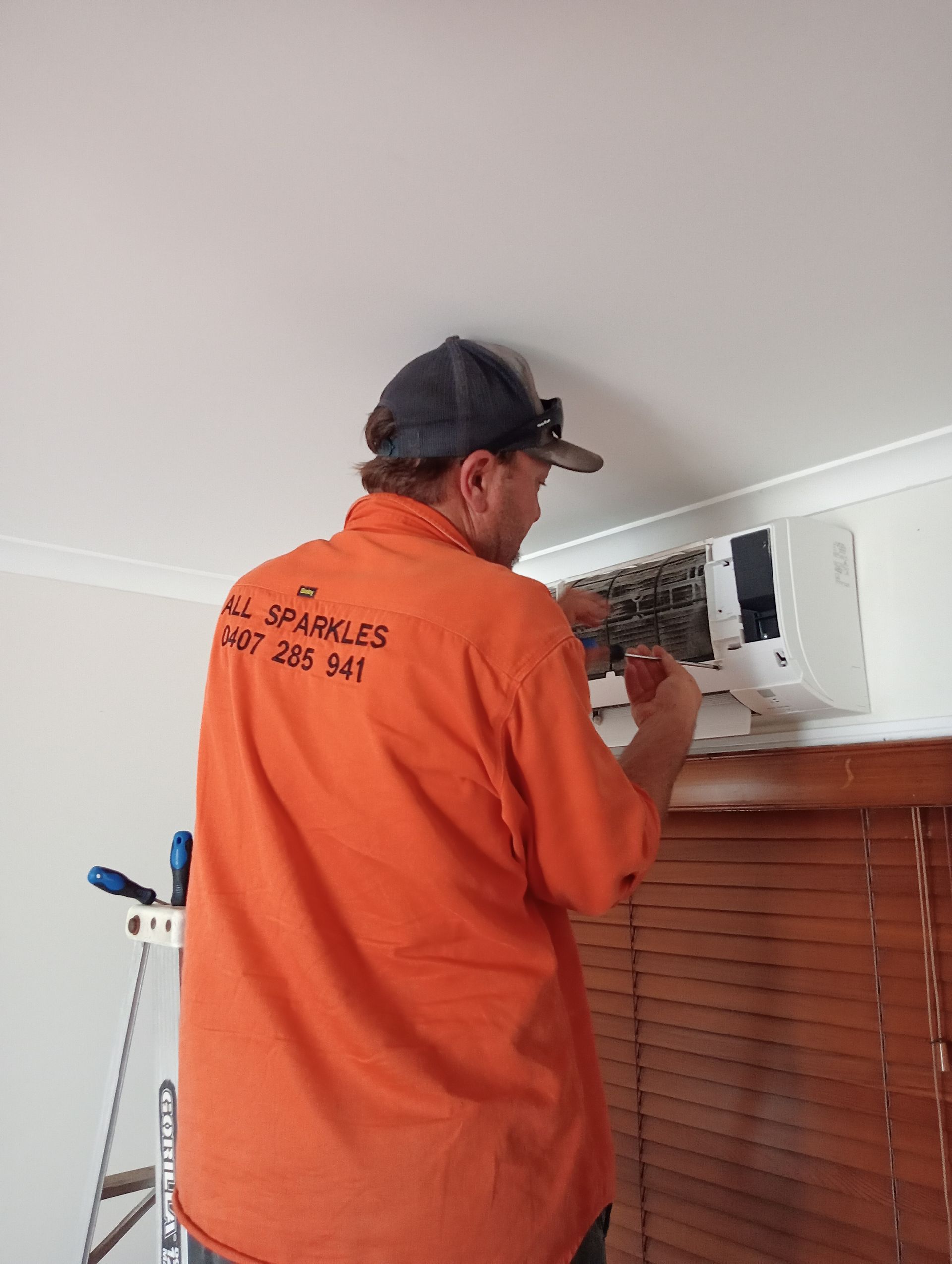 Man in Orange Shirt Servicing an Air Conditioner Mounted on a Wall — All Sparkles Cleaning Technology In Bowen, QLD