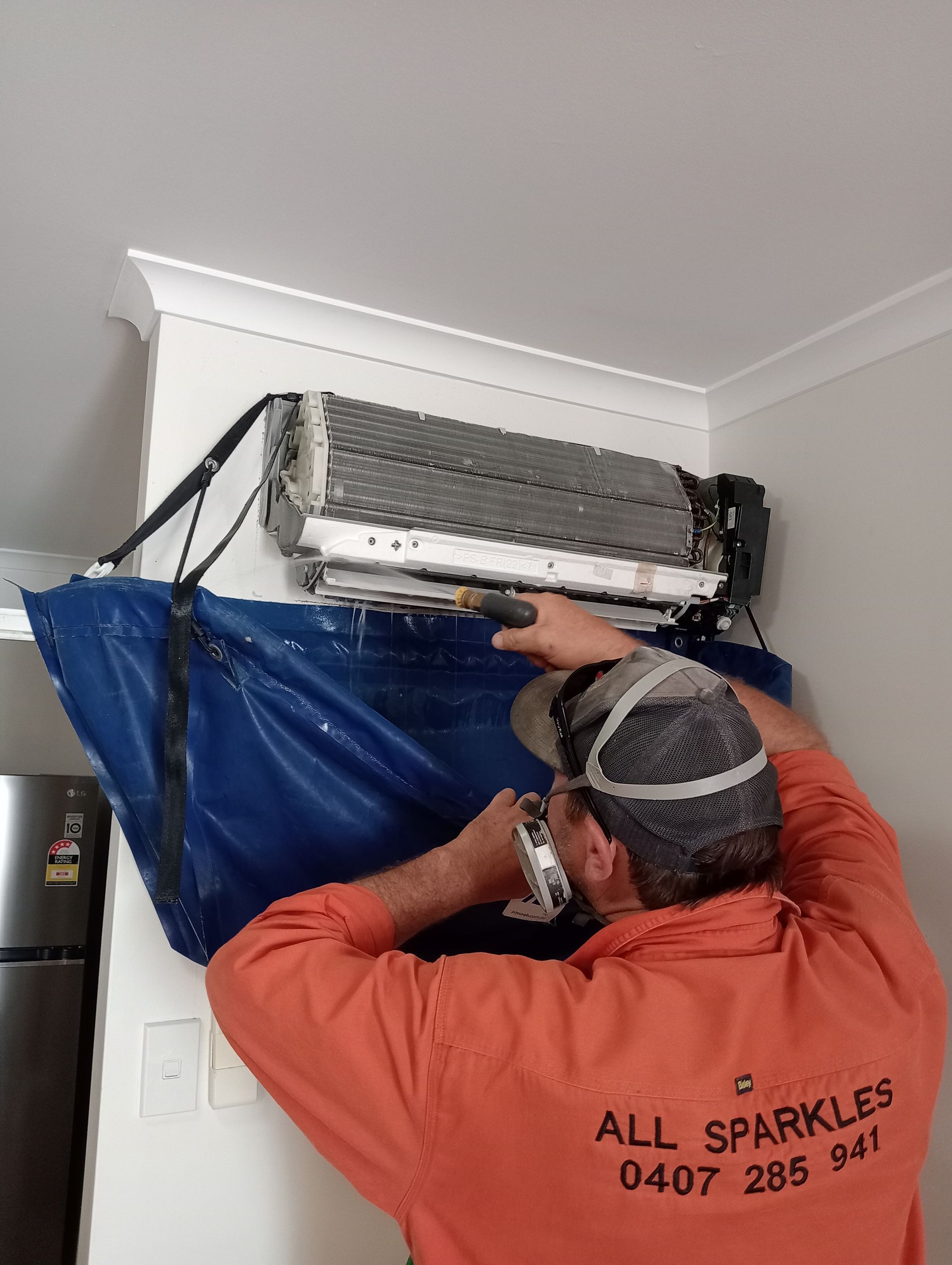 A Person in an Orange Shirt Cleans an Air Conditioner on a Wall — All Sparkles Cleaning Technology In Home Hill, QLD