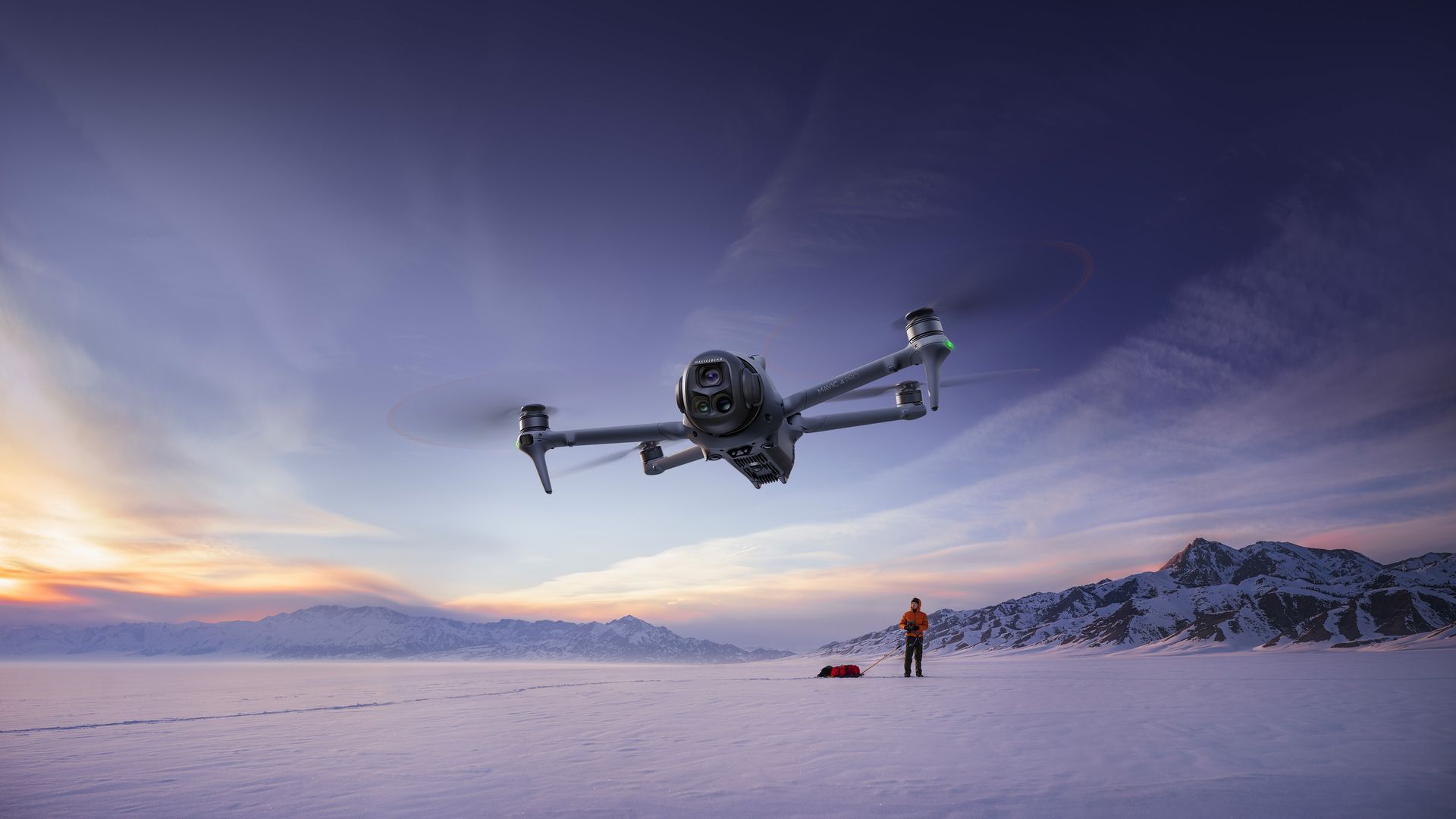 A drone hovers above a vast, snowy landscape at sunset, with a person standing nearby in the distance.