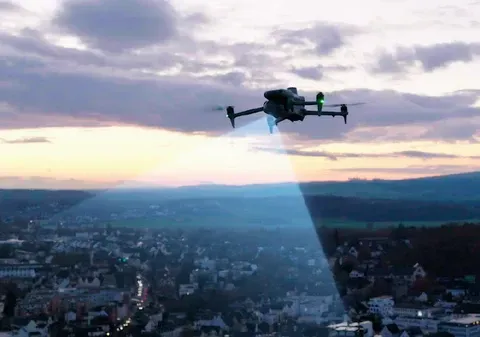 A drone flies over a city at twilight, shining a bright spotlight down onto the buildings below.