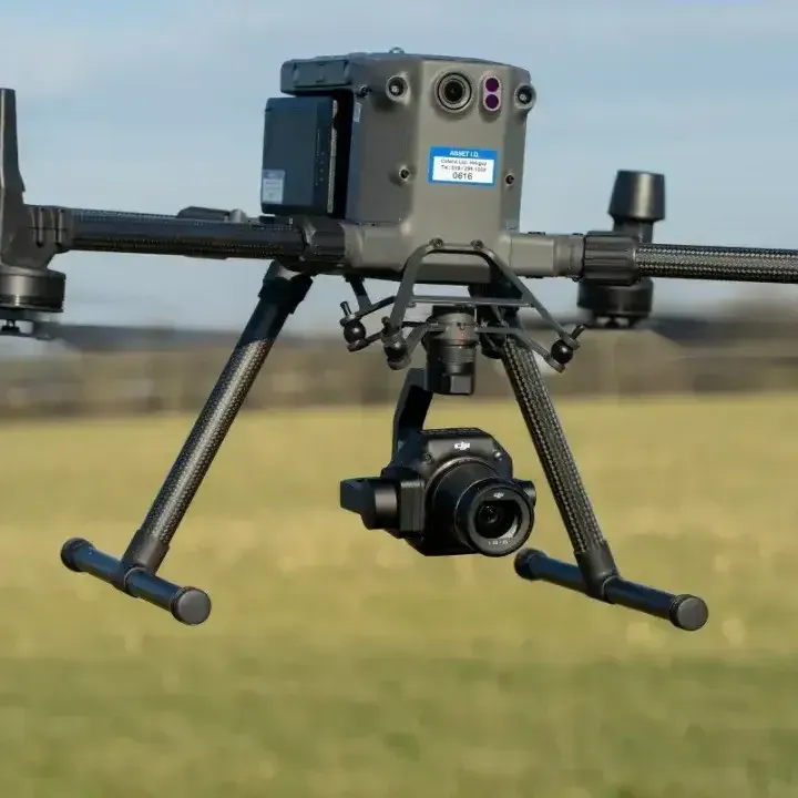 A grey commercial drone hovers in the air above a blurry, golden-green field, featuring a mounted camera and landing gear.