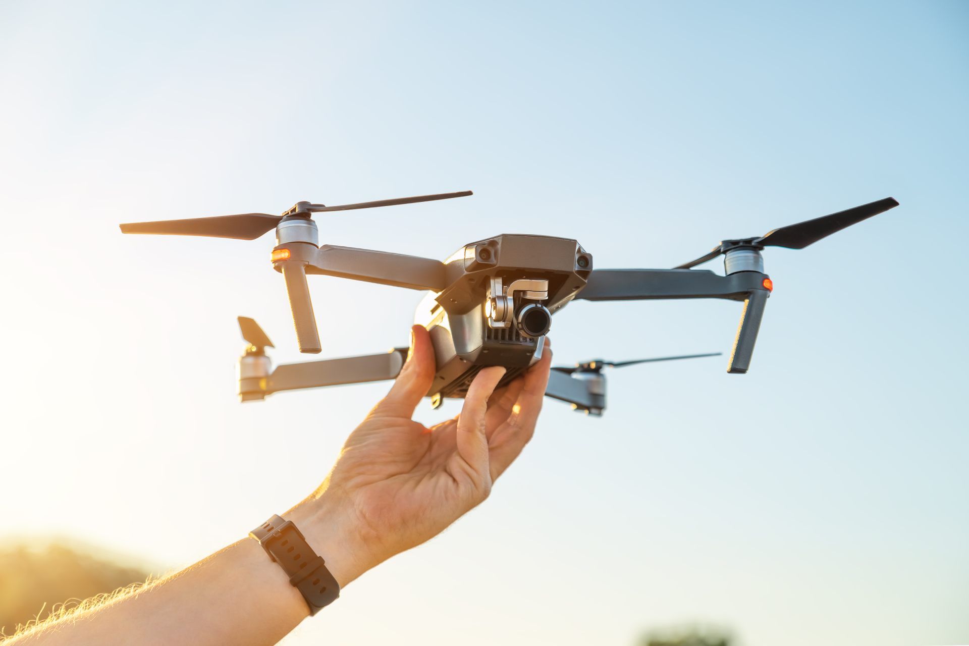 A person holds a grey drone in their hand against a clear blue sky during sunset.