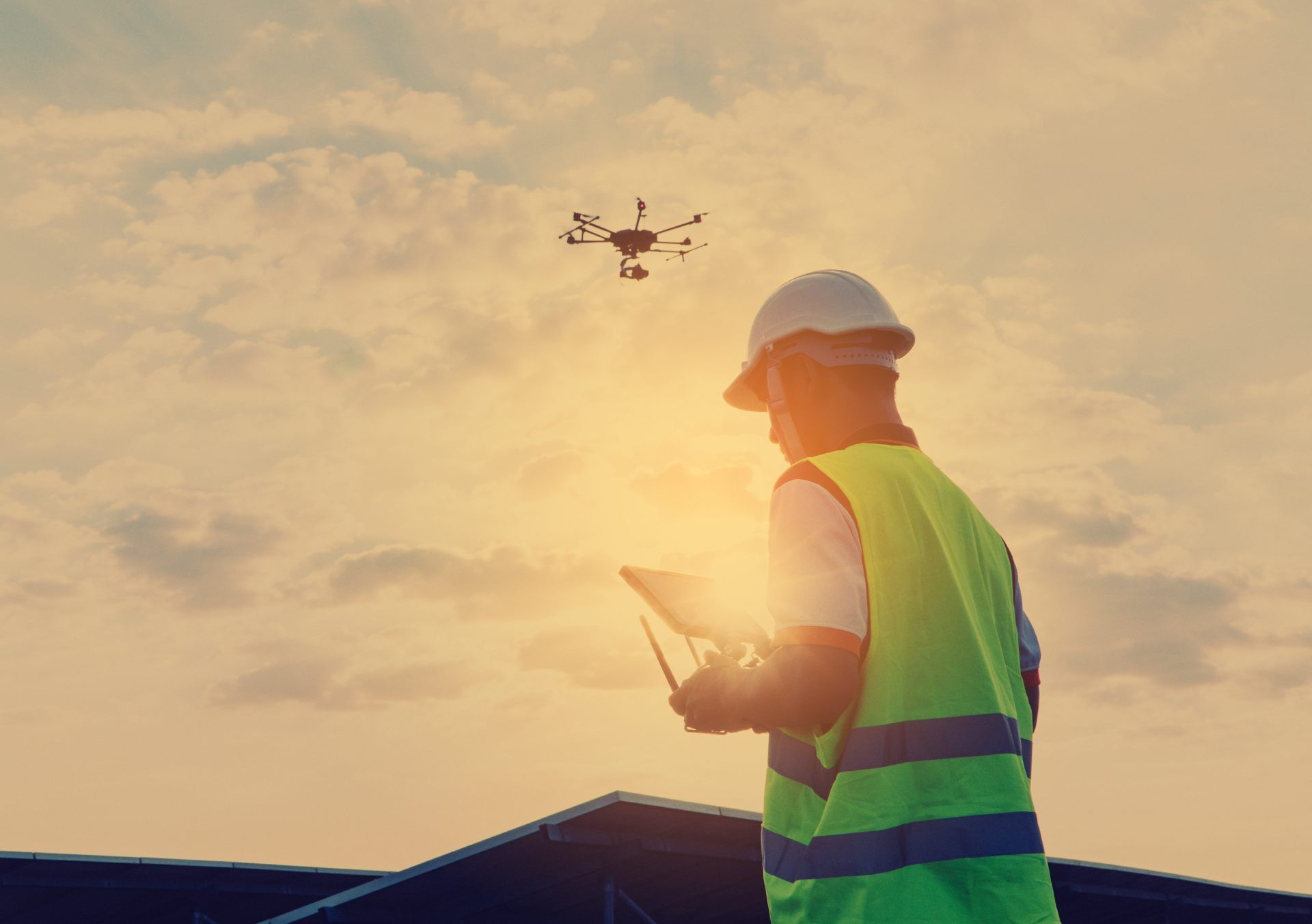 A person operates a drone remote controller during sunset, with a flying drone in the blurred background.