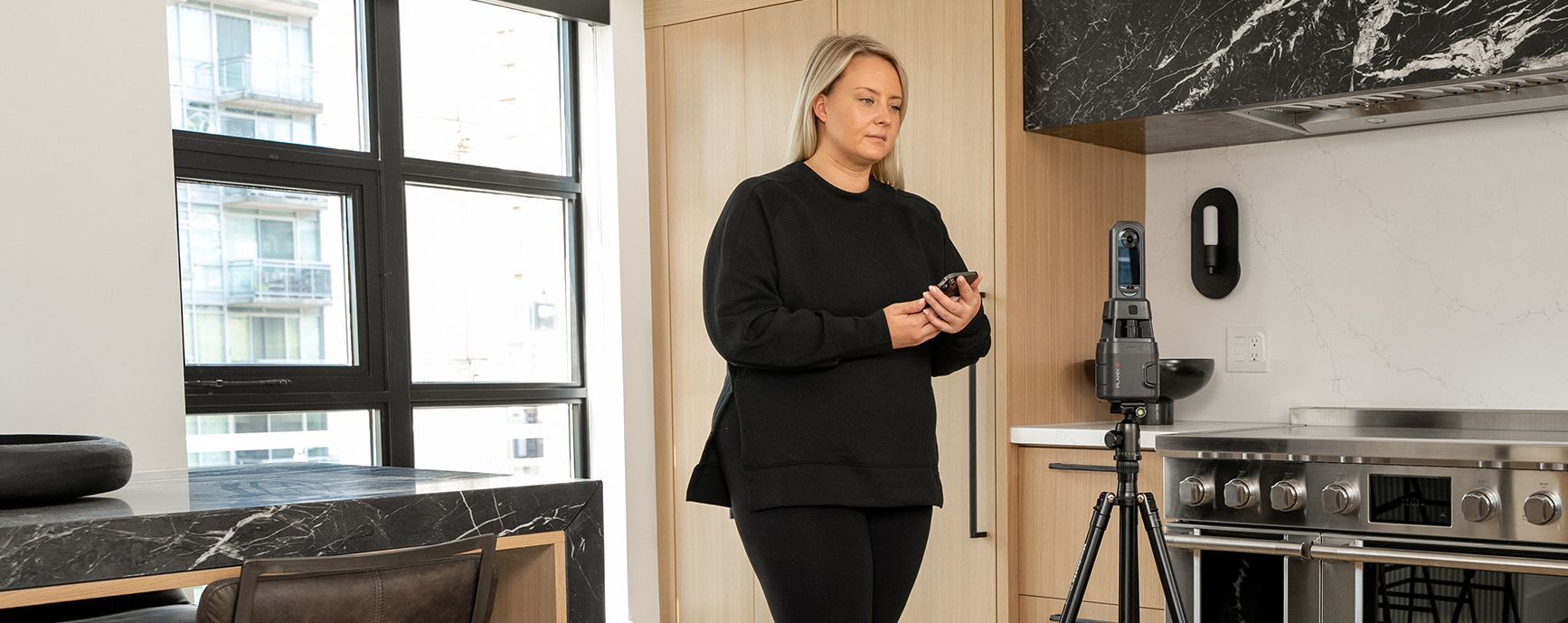 Person standing in a modern kitchen beside a stove and tripod, with large windows behind them