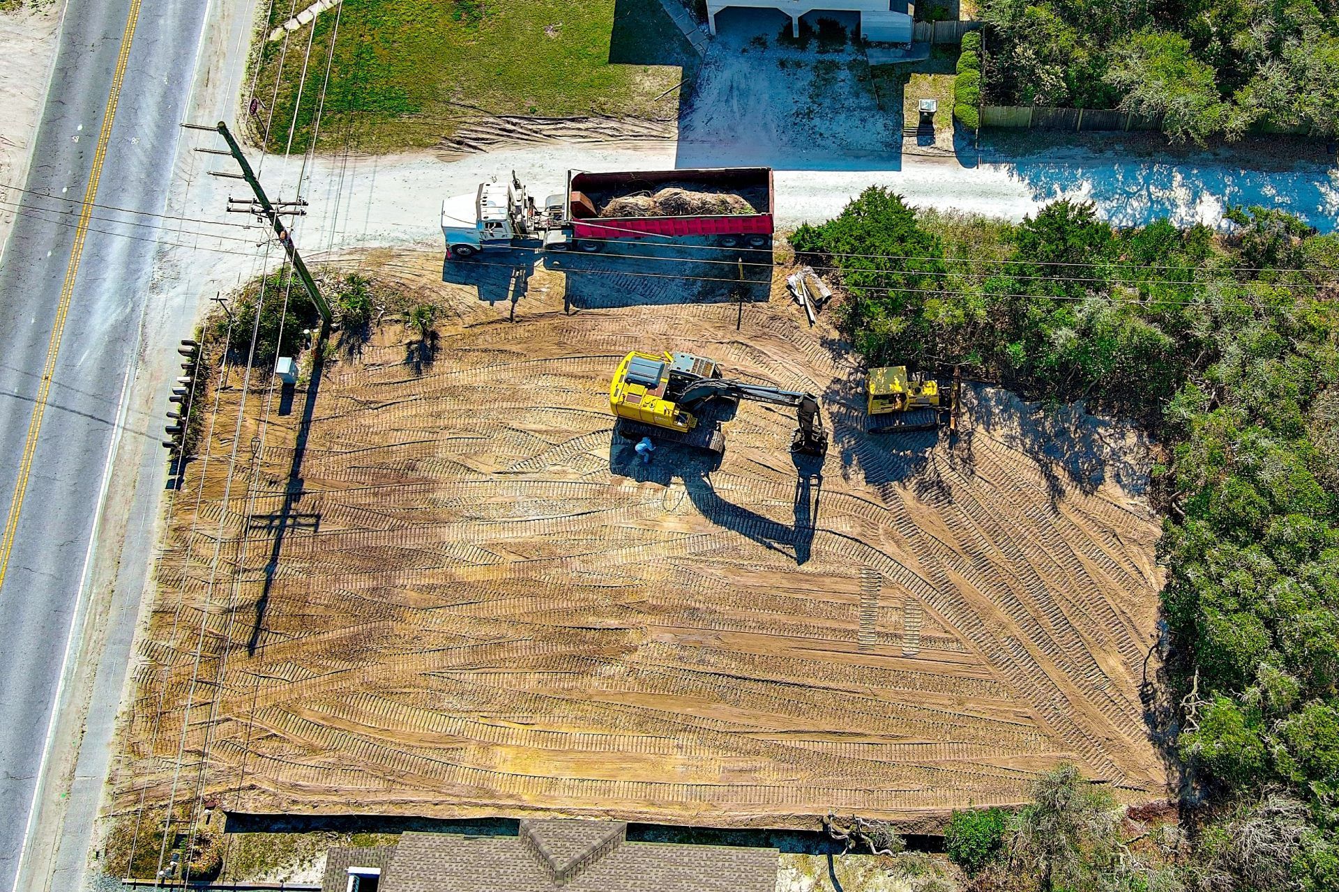 Overhead view of a dirt lot with a small excavator and a red trailer beside a road and trees