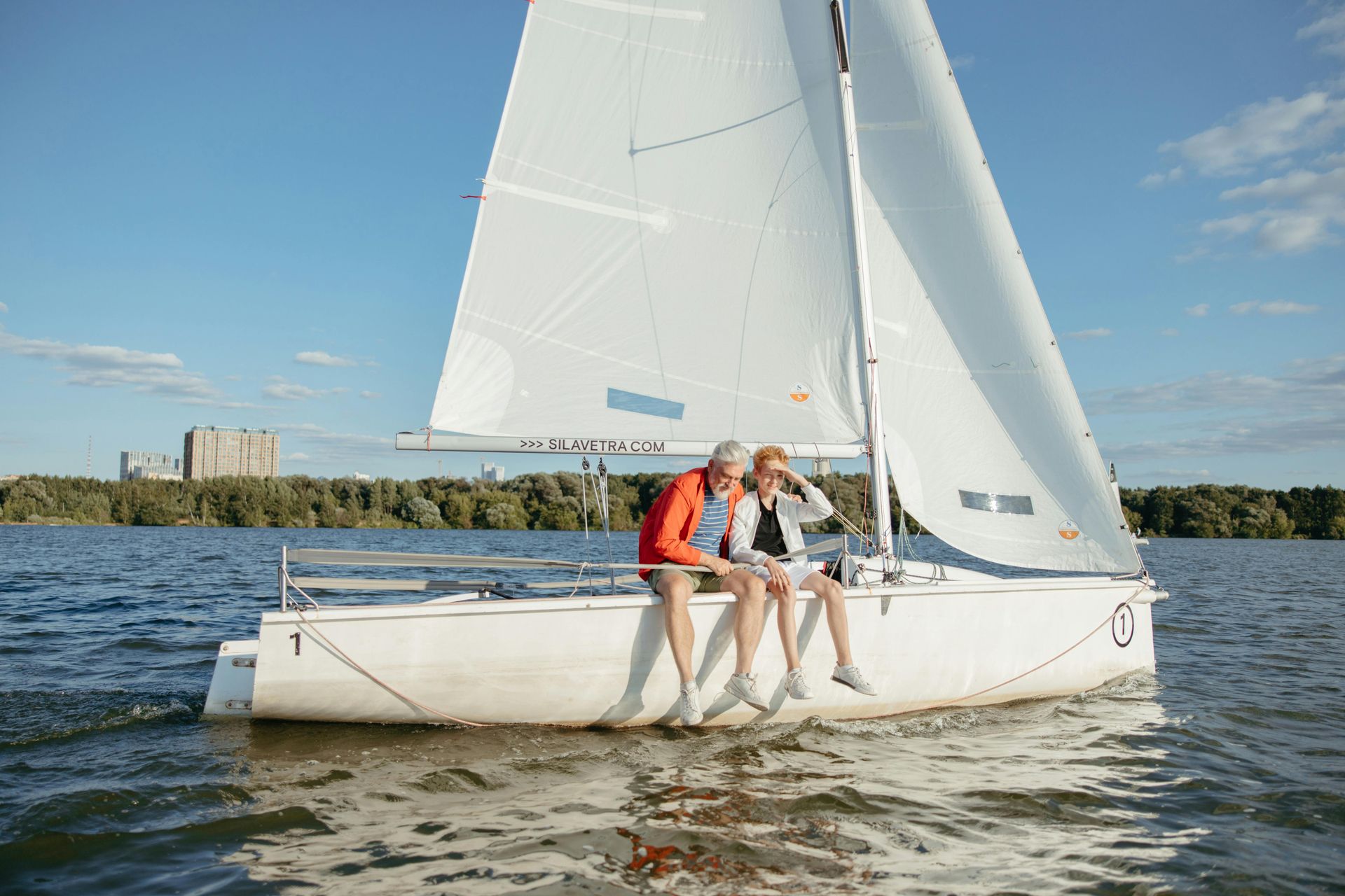 Couple on a sailboat, sitting on the side, white boat on blue water, sunny day.