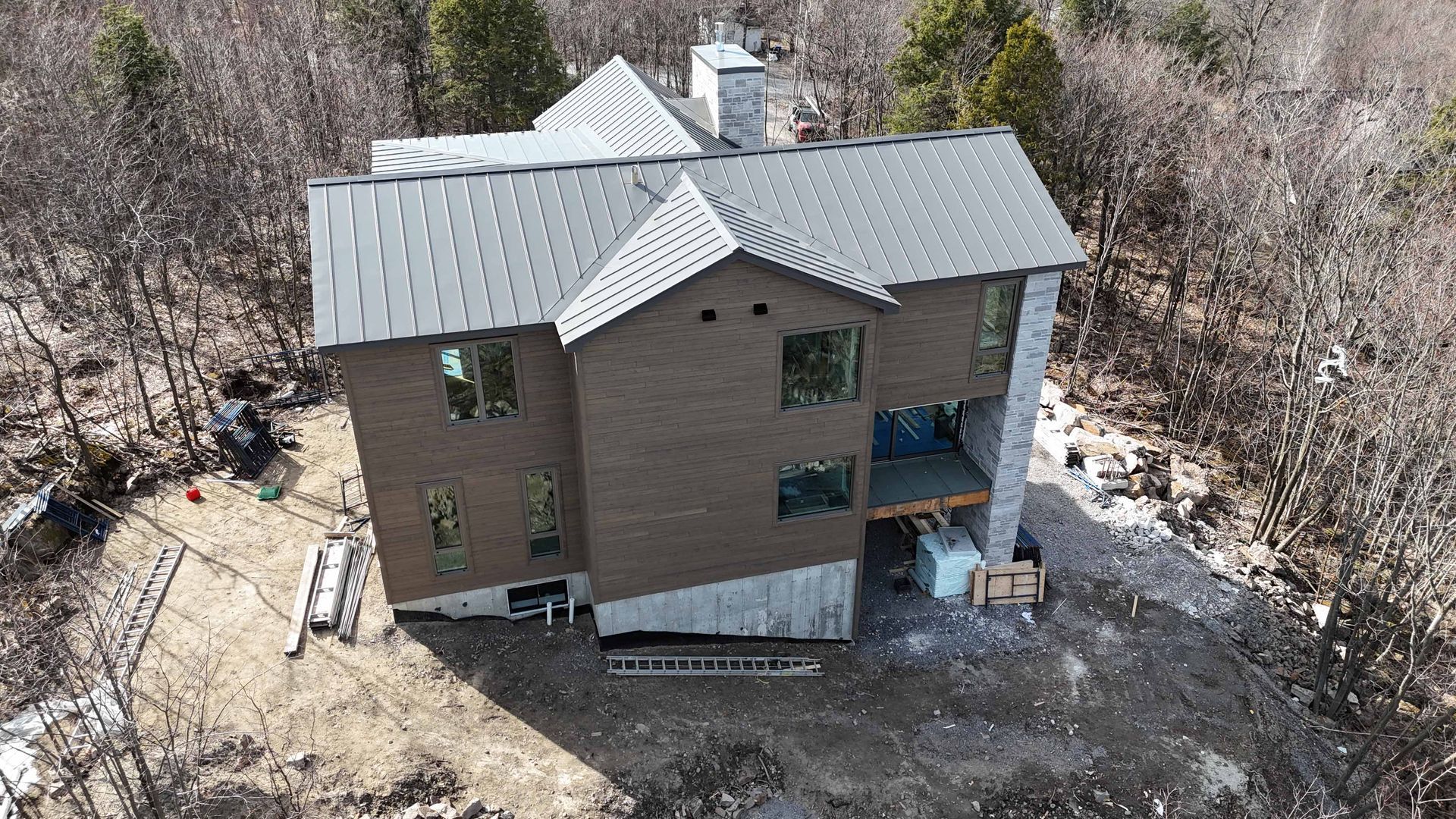 Vue aérienne d'une maison en briques avec un toit en métal, entourée d'arbres dénudés et de plaques de neige.