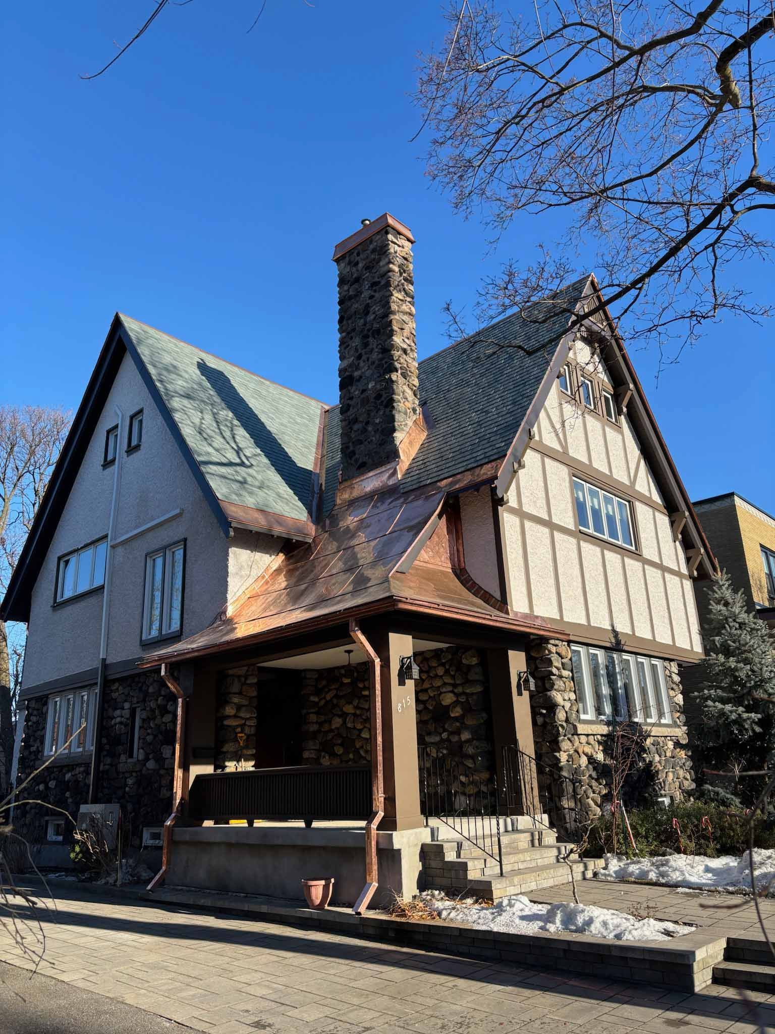 Vieille maison de style Tudor avec pignons abrupts, cheminée en pierre et porche couvert, située dans une rue ensoleillée.