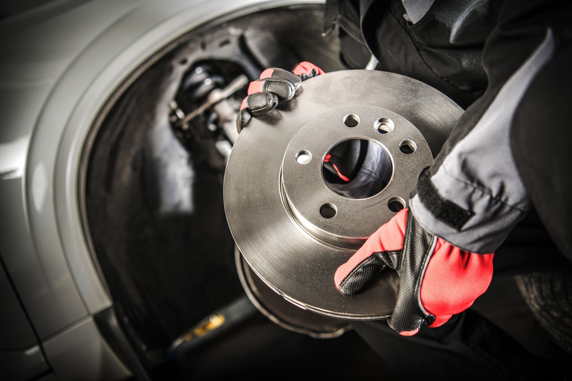 AC scrolling— A/C Service in Daytona Beach, FL Gloved hands holding new brake disc beside exposed car wheel and brake assembly.