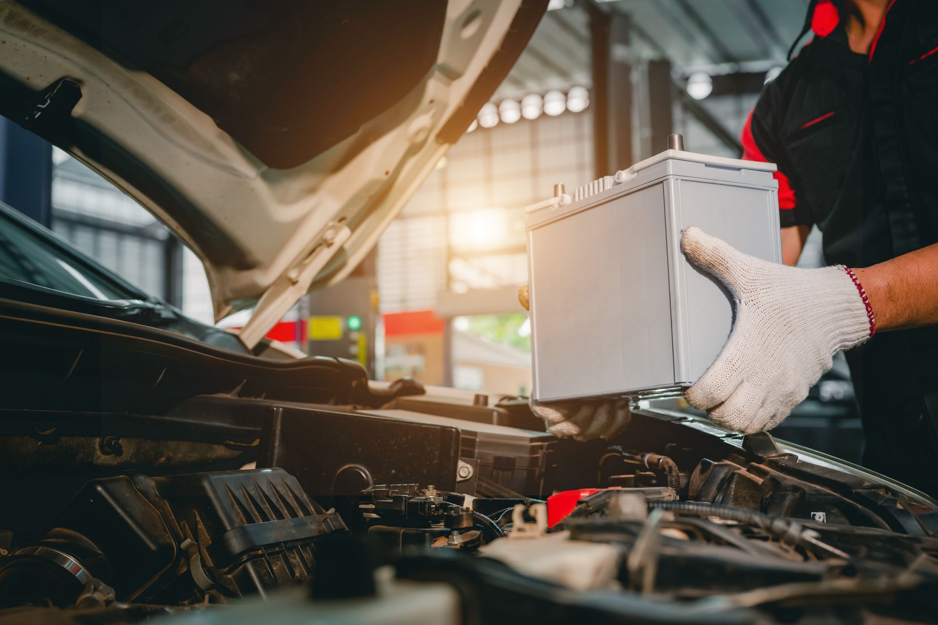 Mechanic installing a car battery under an open hood in an auto repair shop.