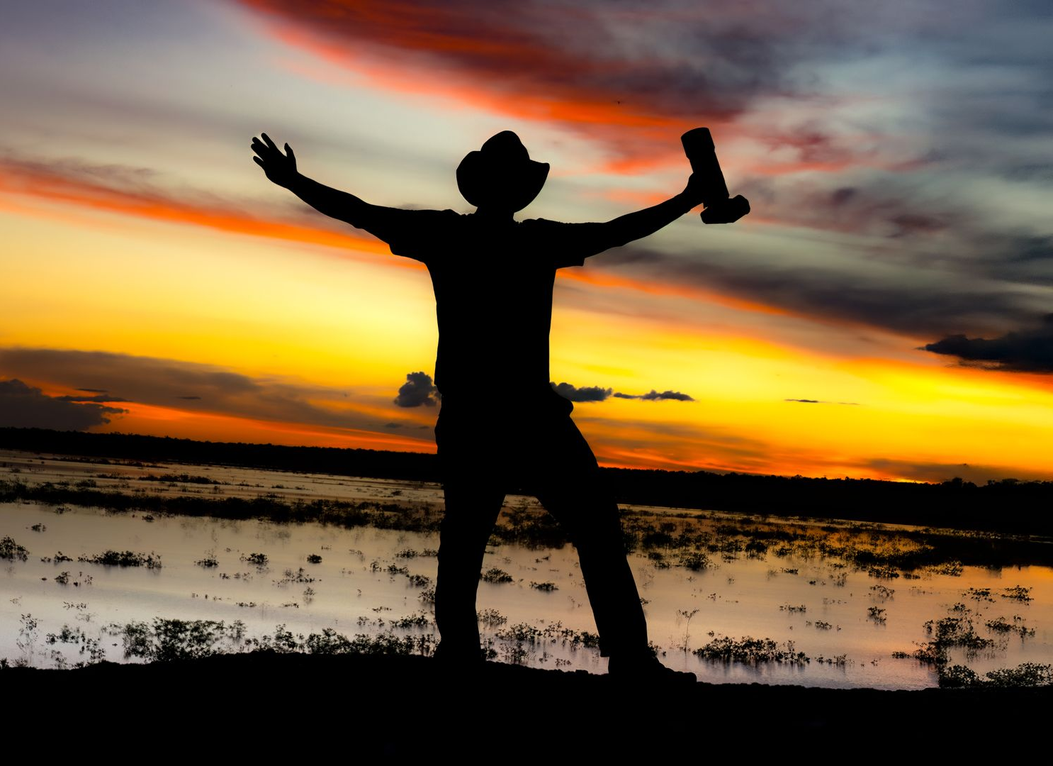 Silhouetted person with arms raised at sunset beside a calm body of water