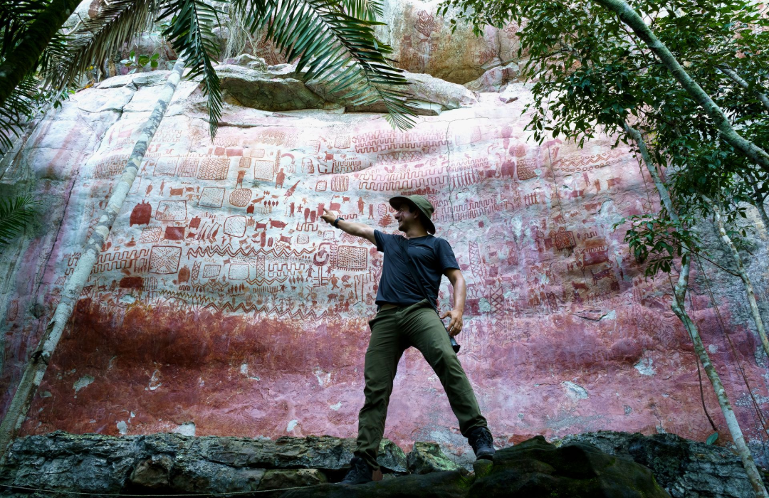 Guide standing on rocks beneath a large red-and-white cliff, pointing upward among palm fronds.