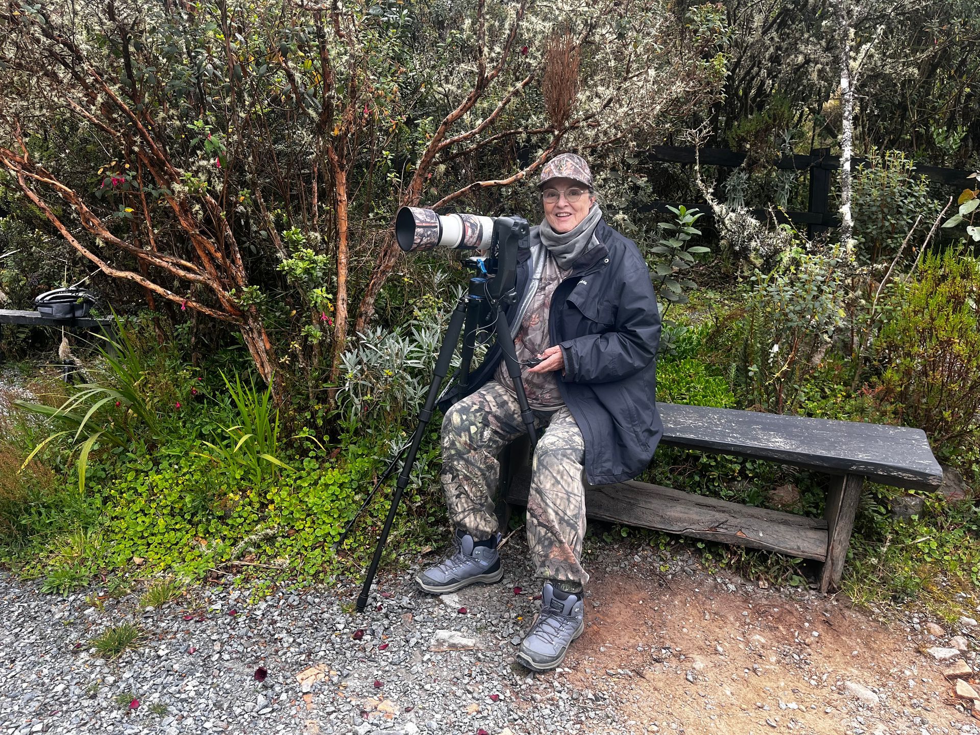 Person sitting on a bench outdoors, holding a large telephoto camera in a wooded area.