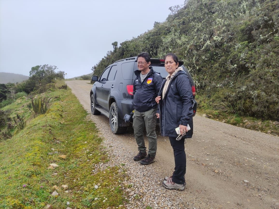 Two Bird photographers stand beside a dark SUV on a gravel mountain road with green hills in the.